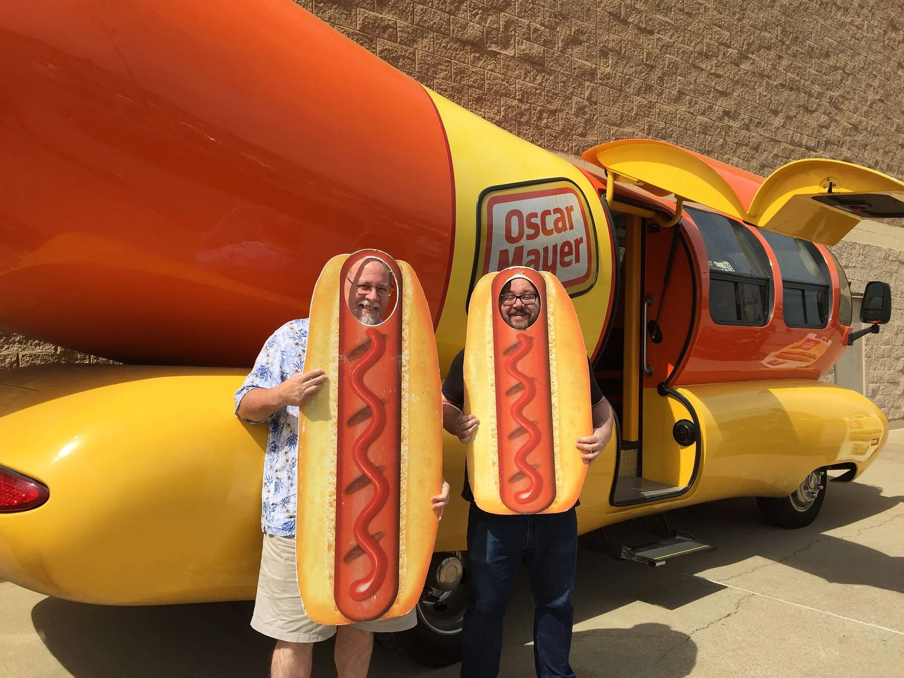 Two men posing with the Oscar Mayer Wienermobile and hot dog cutouts.