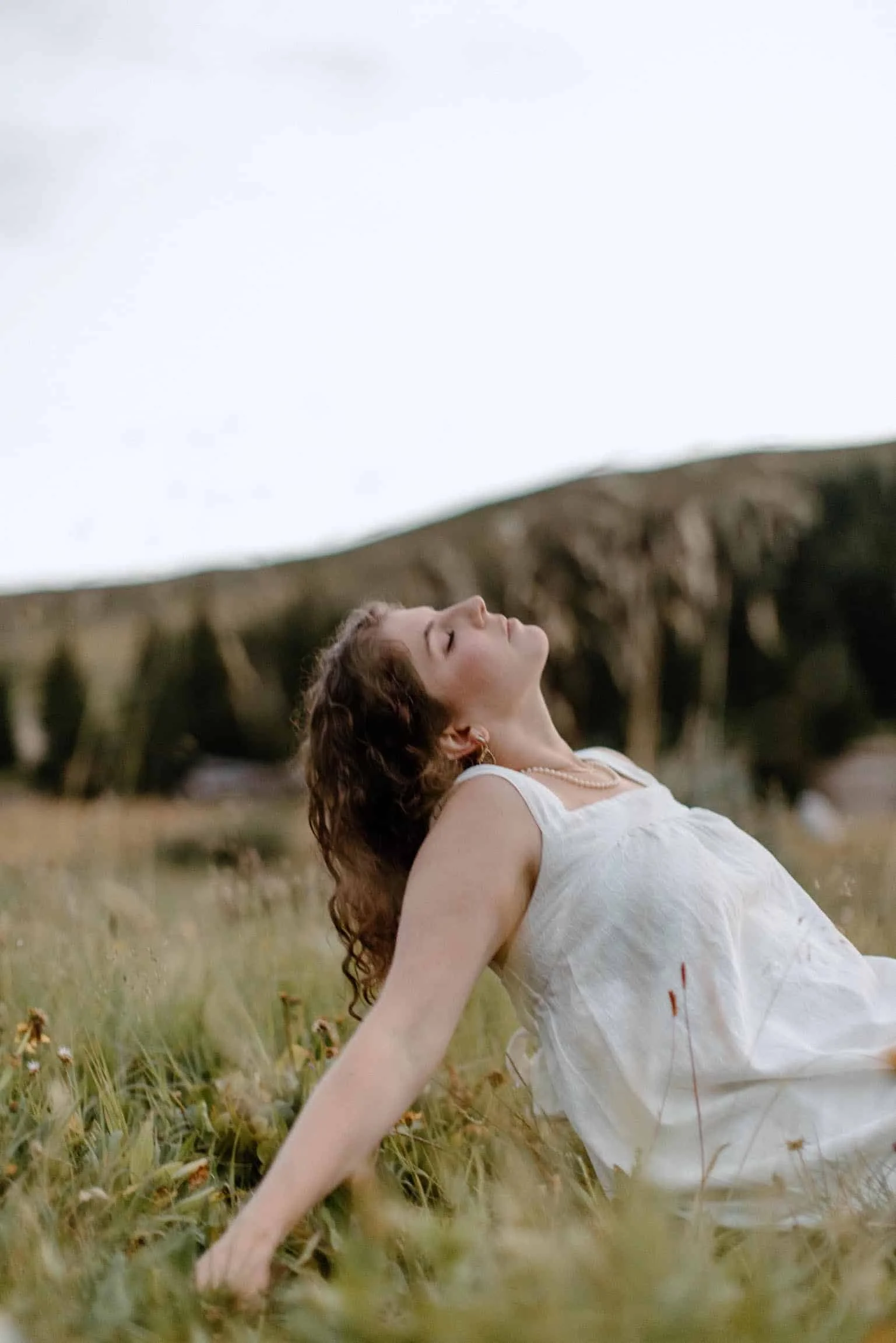 brunette woman in flowing white tank top sits on grass and leans back onto outstretched arms with her eyes closed