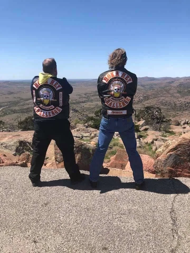 Two men wearing motorcycle jackets with their backs to the camera, looking out over a desert.