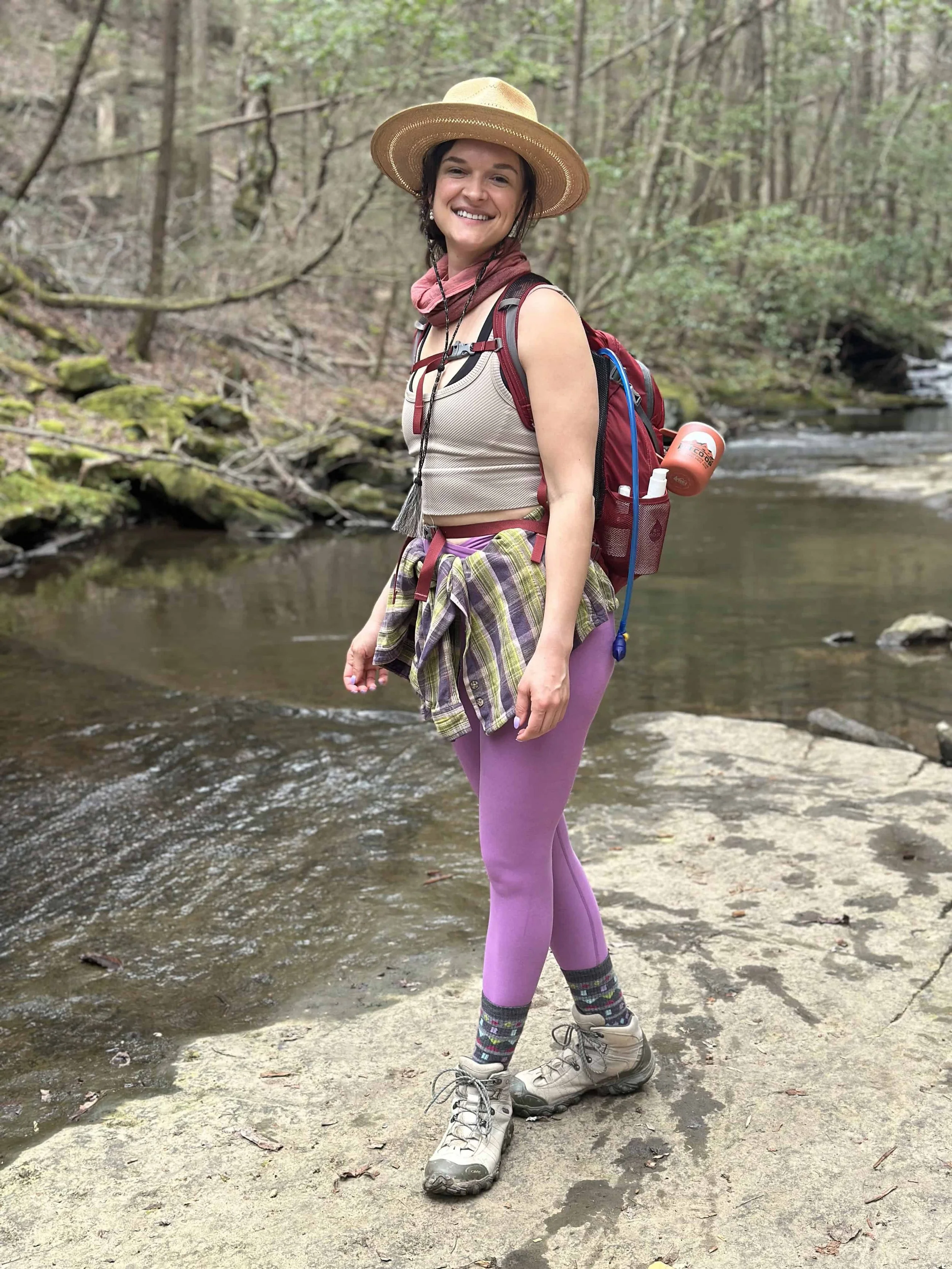 Woman in a hat standing on a rock next to a creek during a hike in the woods.