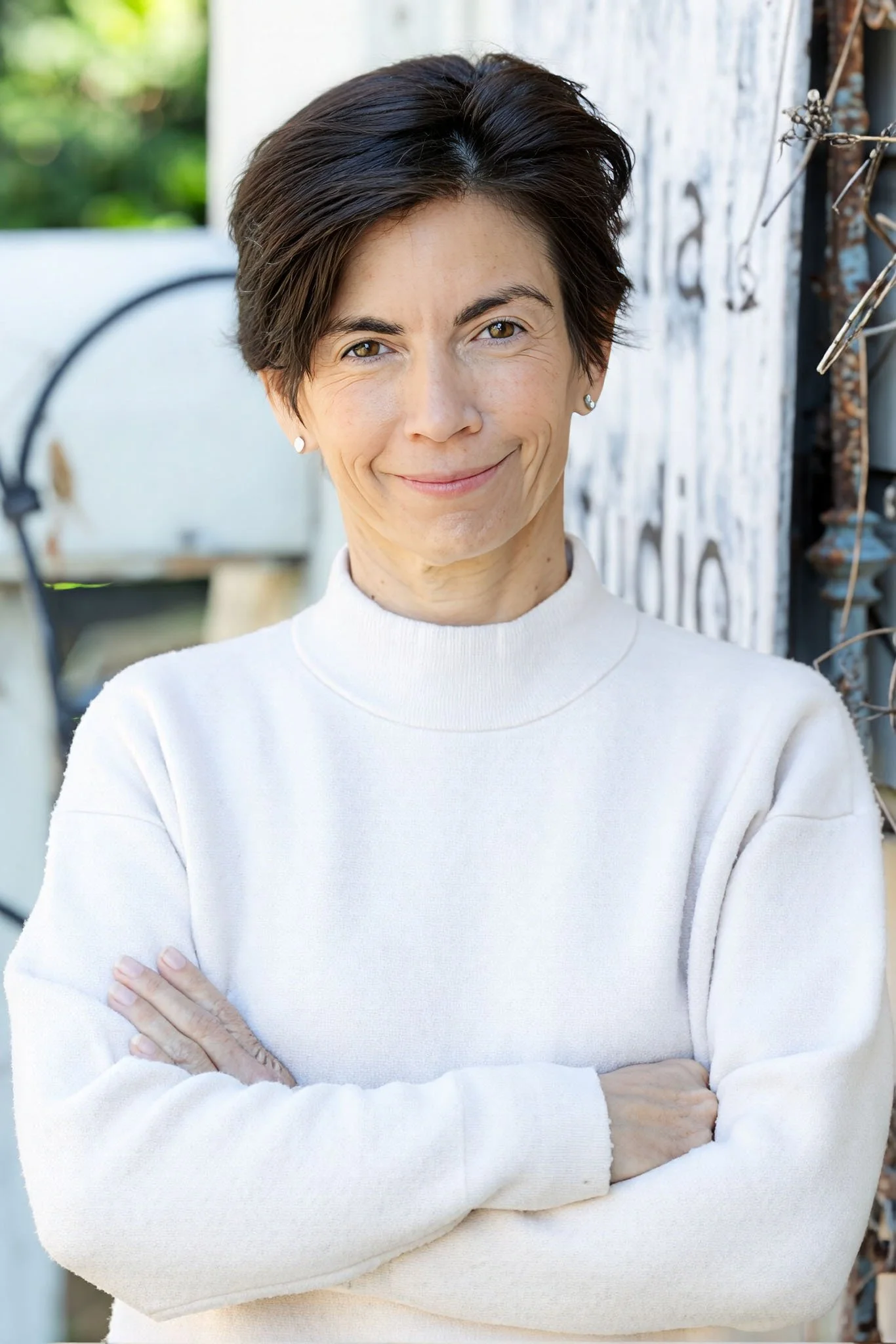 Woman with short dark hair wearing a white sweater, standing outdoors with her arms crossed and smiling at the camera.