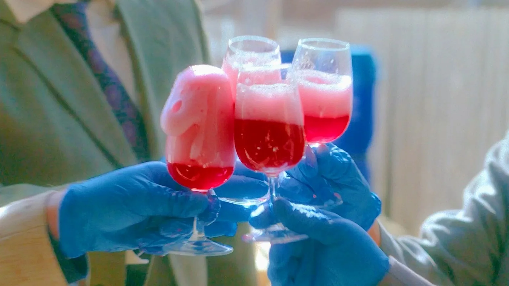 Three glasses of pink sparkling beverage with frothy foam, held by two people wearing blue gloves.