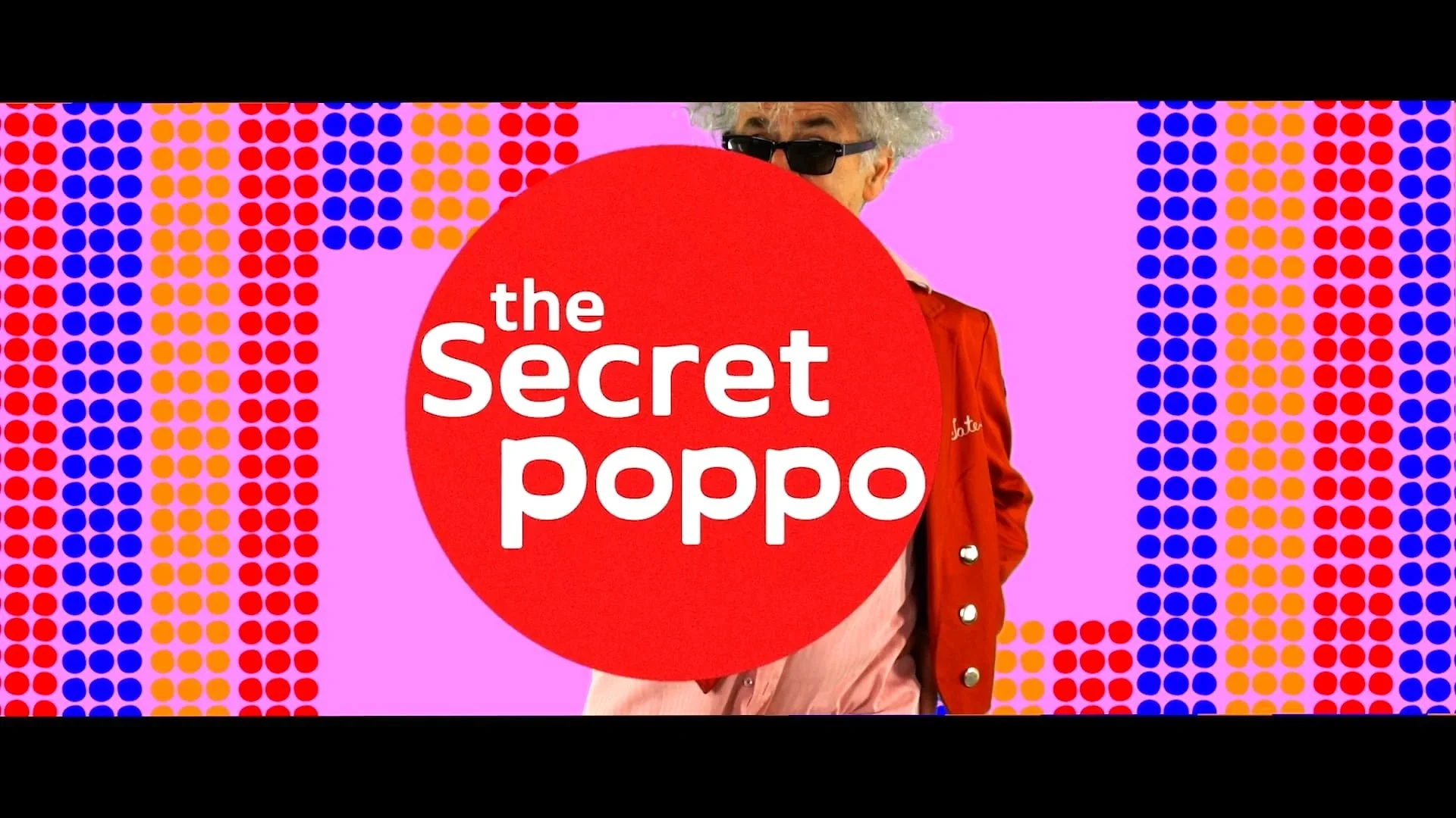 A person with white, curly hair and sunglasses, standing behind a large red circle with the text 'the secret poppo' on it. The background features a colorful pattern of blue, red, orange, and purple dots on a pink backdrop.