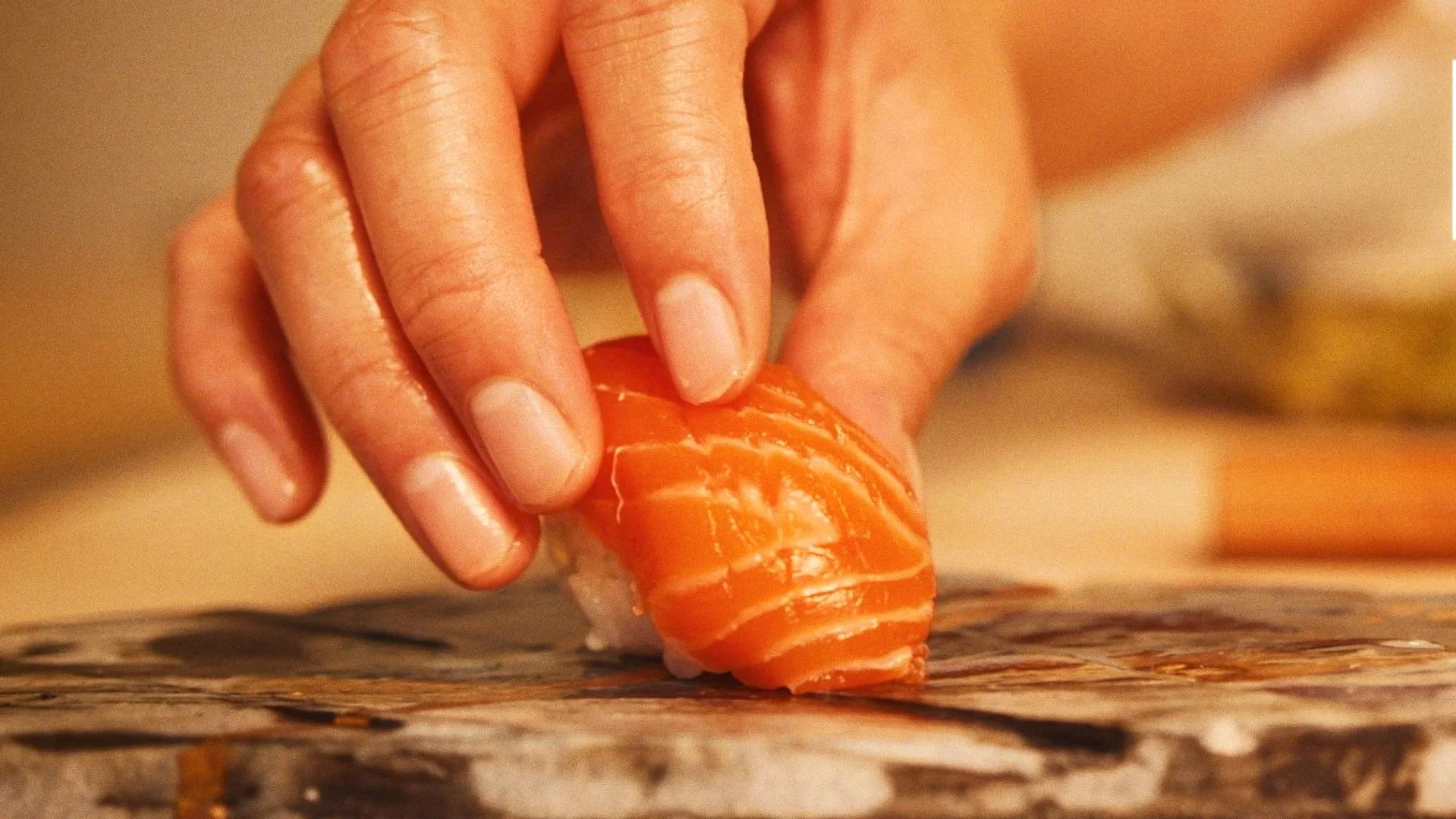 Close-up of a hand placing a piece of salmon on a piece of sushi on a surface.