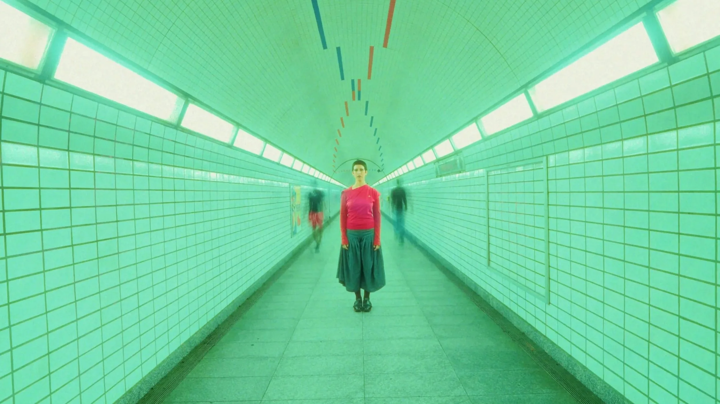 A woman in a red top and long skirt standing in a green-tiled tunnel, with blurred figures walking behind her.