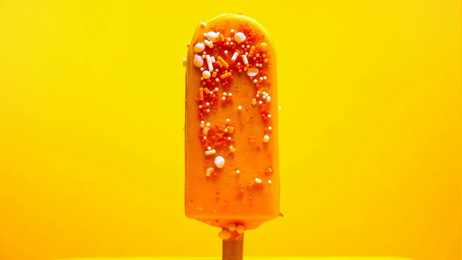 Orange popsicle on wooden stick decorated with colorful sprinkles against a yellow background.