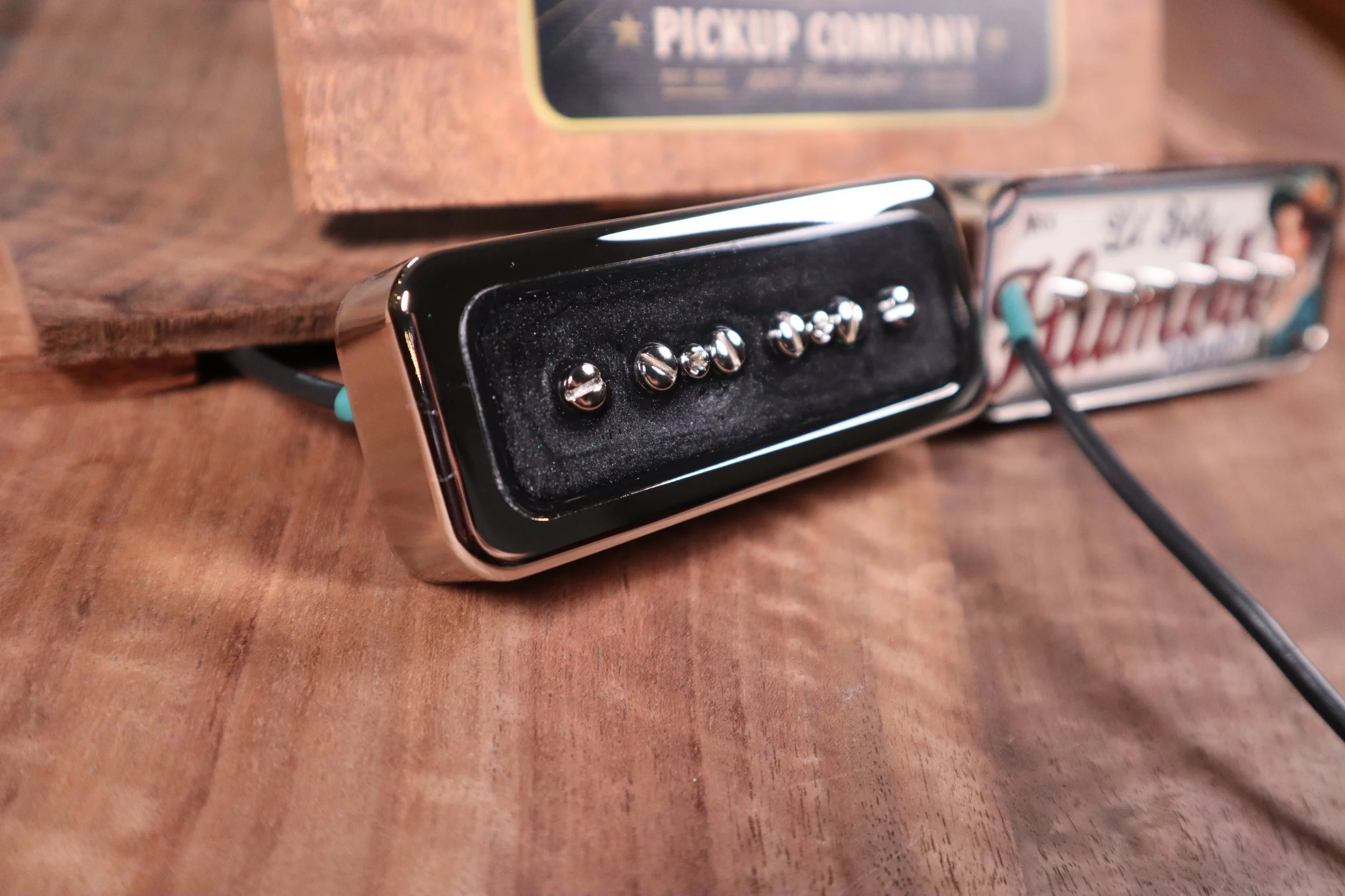 Close-up of a black guitar P-90 Lil' Belle pickup on a wooden surface with a partially visible box in the background.