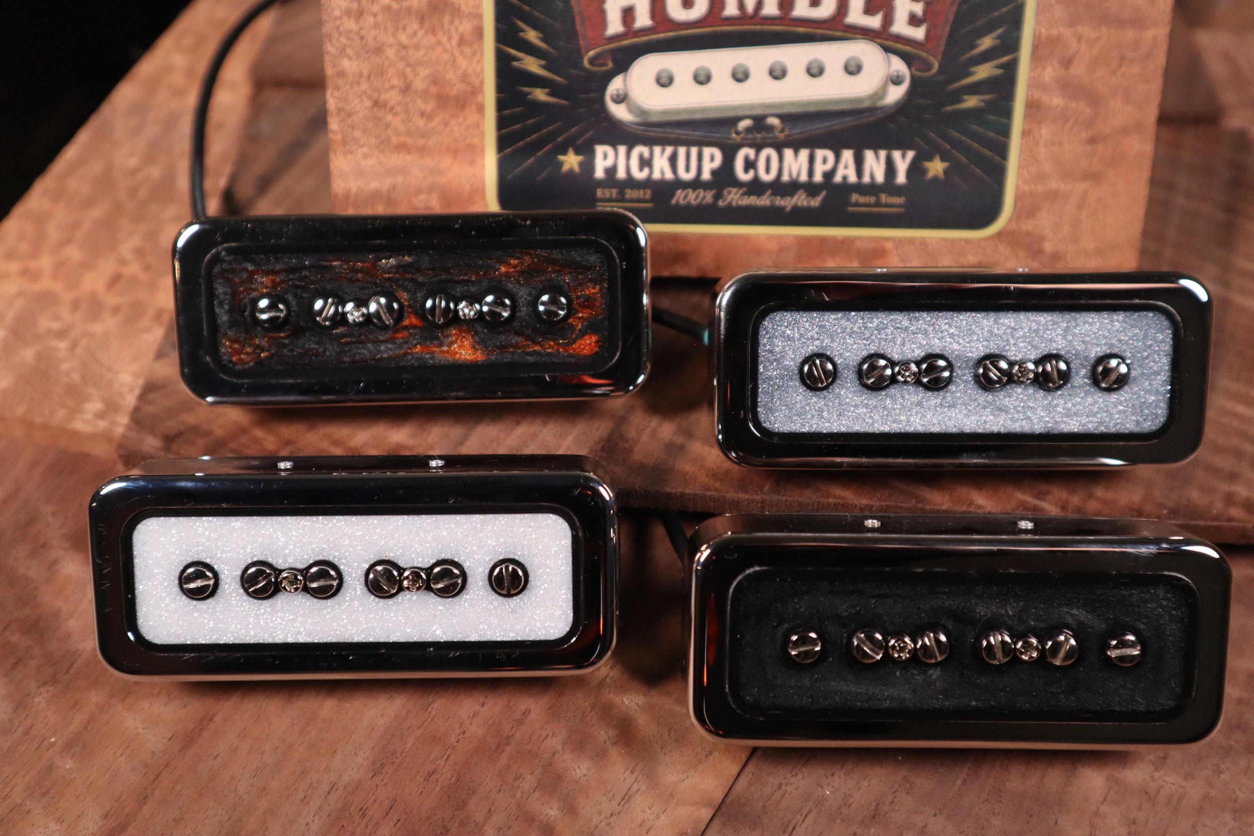 Close-up of several guitar pickups placed on a wooden surface, with a box labeled 'Humble Pickup Company' in the background.
