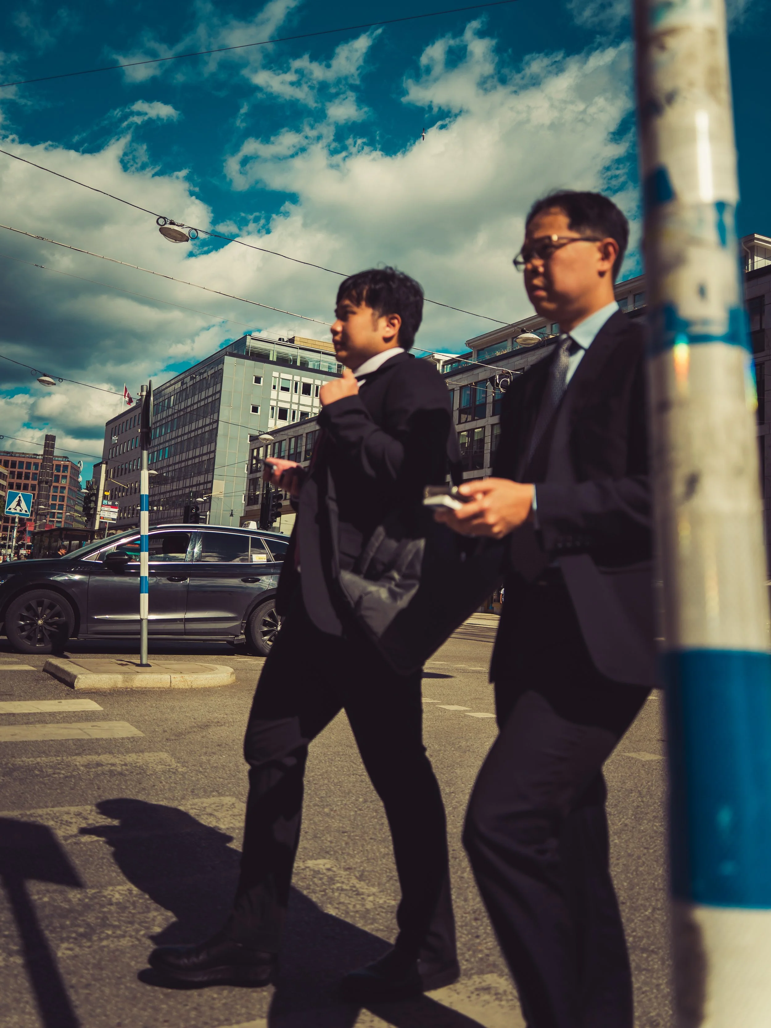Two men in suits walking across a city street, one looking at a phone and the other adjusting his collar, with cars and buildings in the background and a cloudy sky overhead.