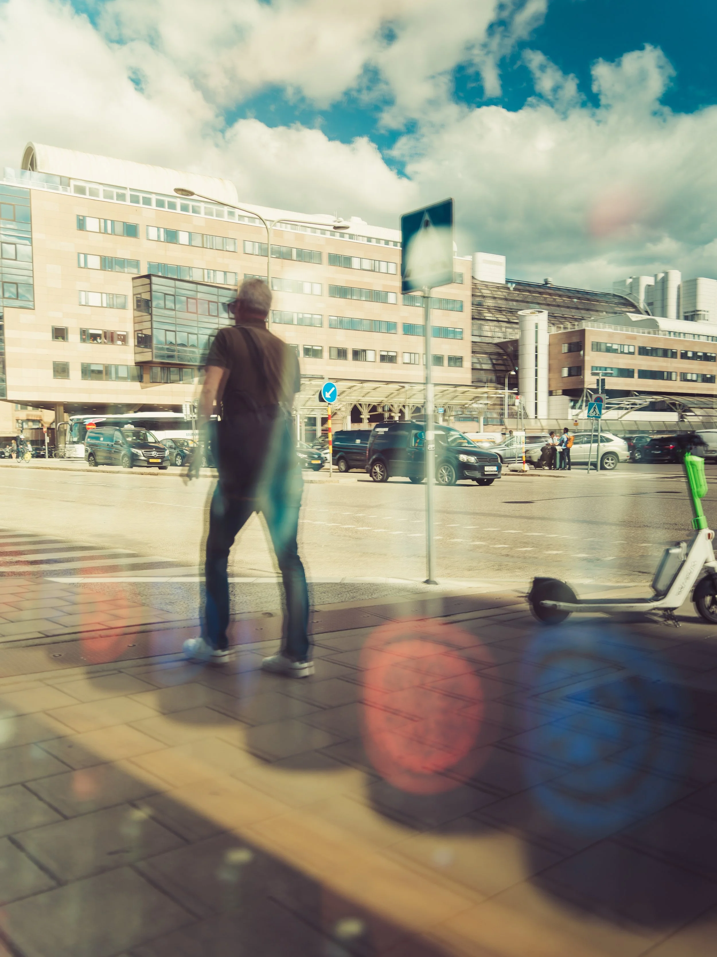 A person walking on a city sidewalk near parked cars and a scooter, with a modern building and cloudy sky in the background.
