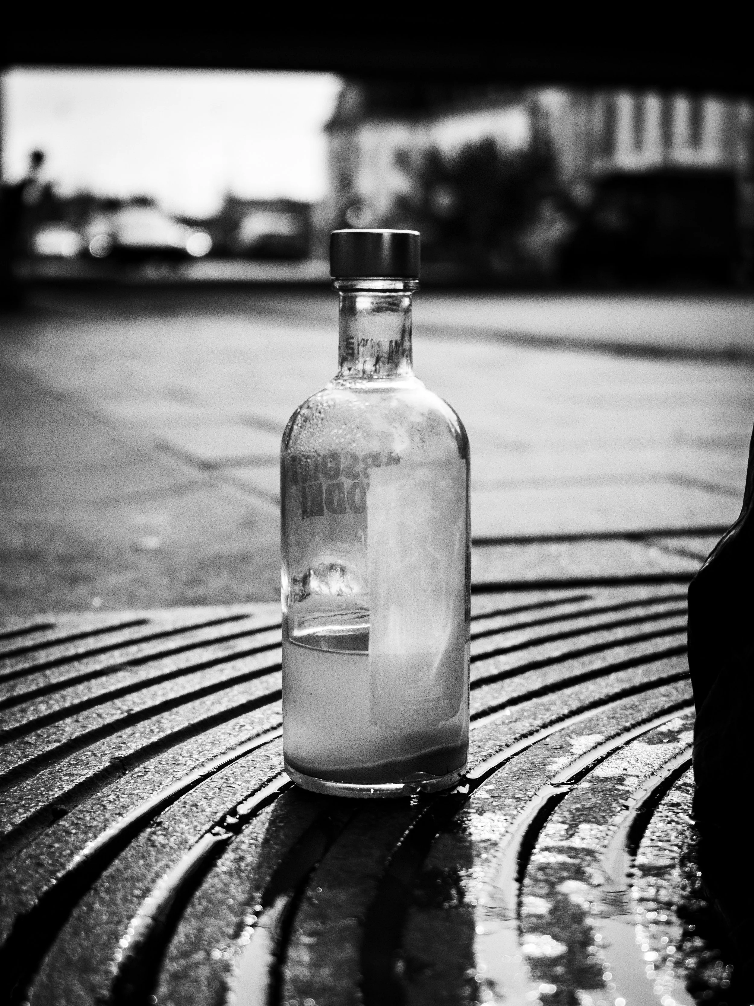 A black and white photo of a glass bottle placed on a wet, textured surface with parallel grooves. The bottle appears to have a liquid inside and is capped. Blurred background with cars and buildings, possibly taken outdoors in an urban setting.