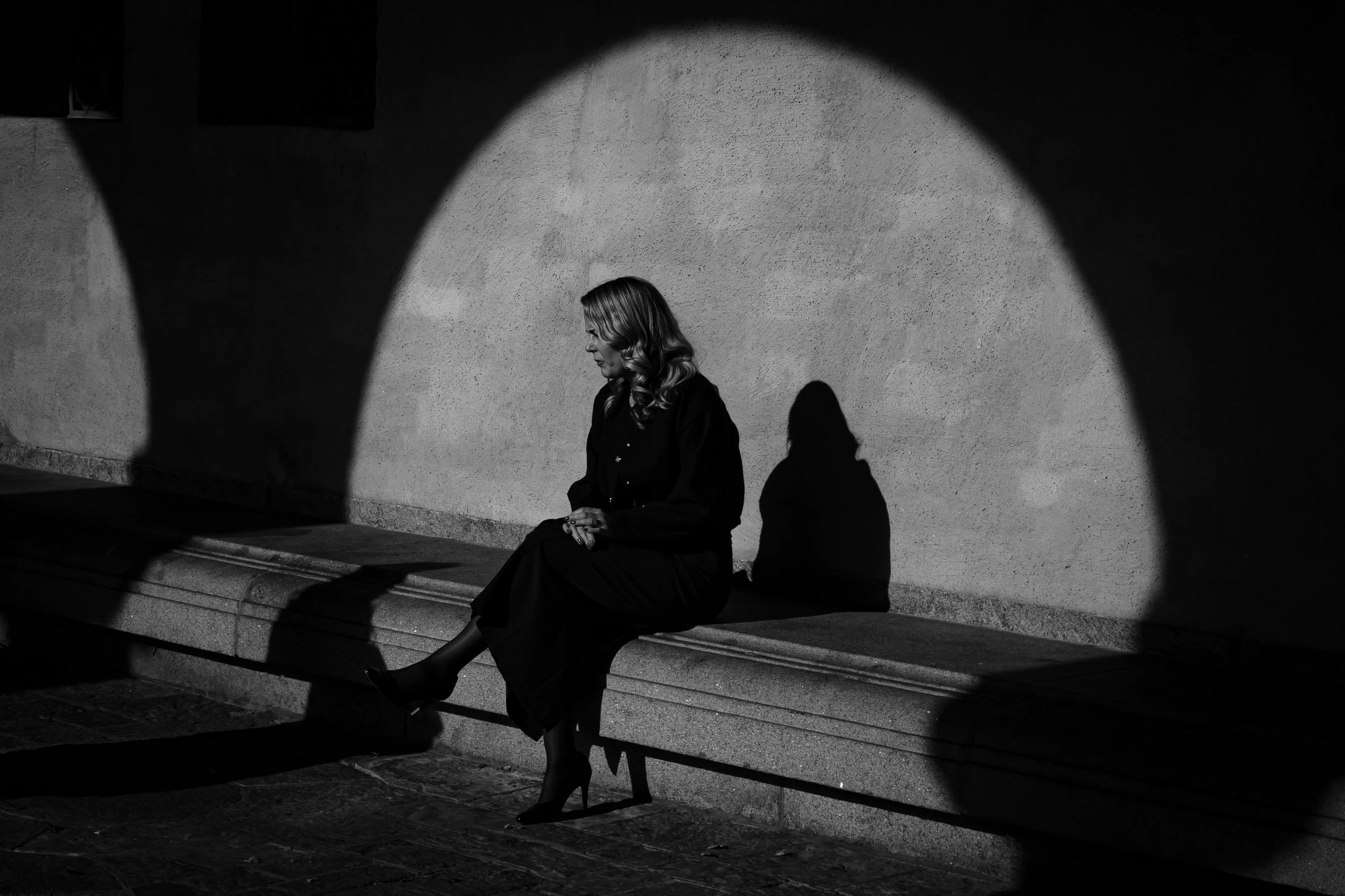 A woman sitting on a bench in a shadowed area with a wall behind her, illuminated by a spotlight, creating a shadow on the wall.