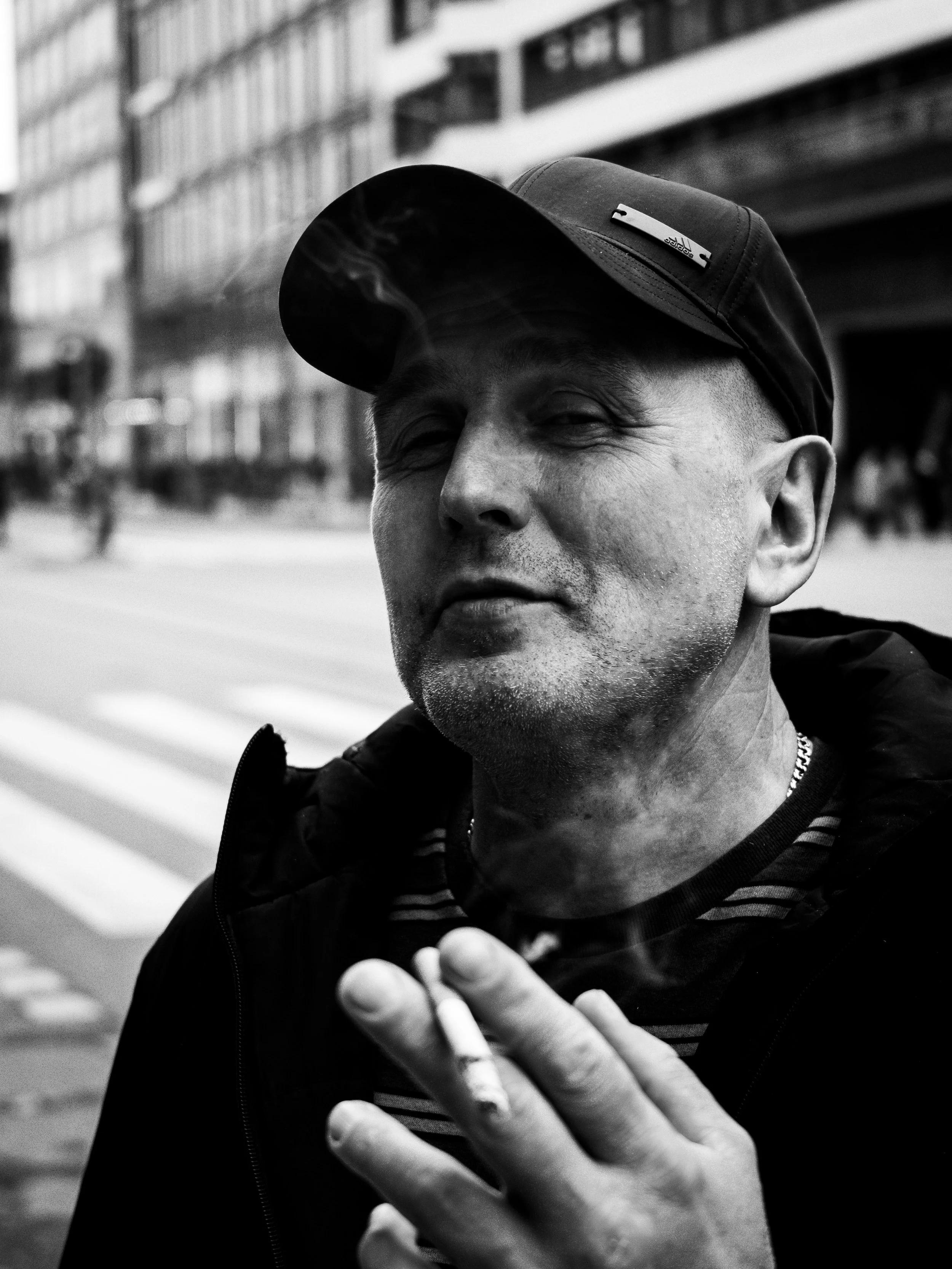 A middle-aged man with short hair and a beard, wearing a baseball cap and striped shirt, smokes a cigarette on the street in an urban area.