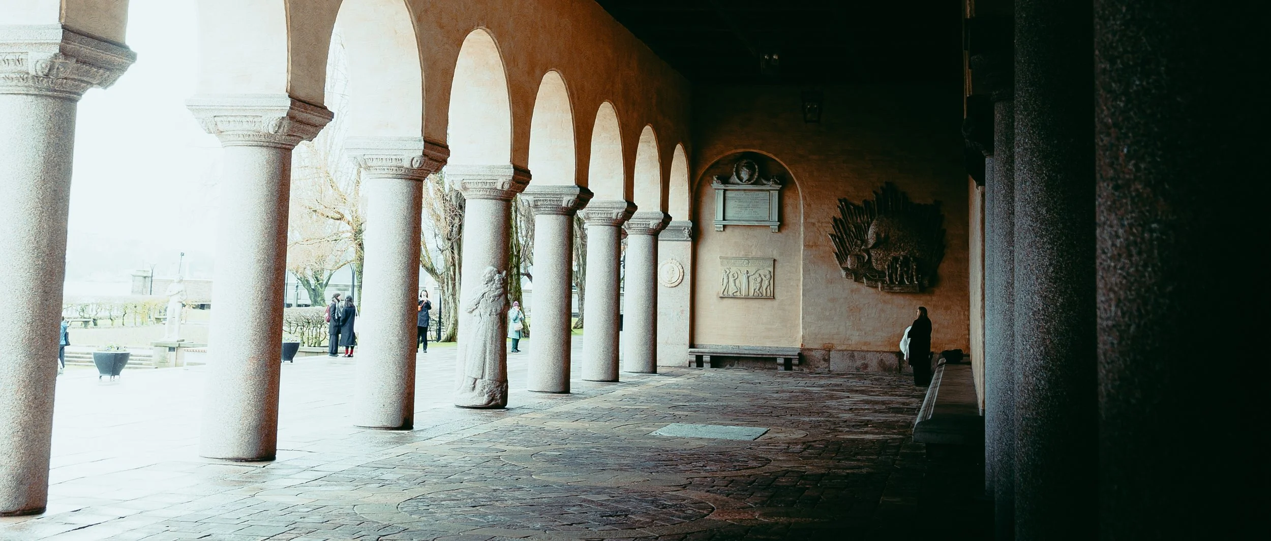 Arcade with stone columns and sculptures inside, overlooking an outdoor area with people walking and trees.