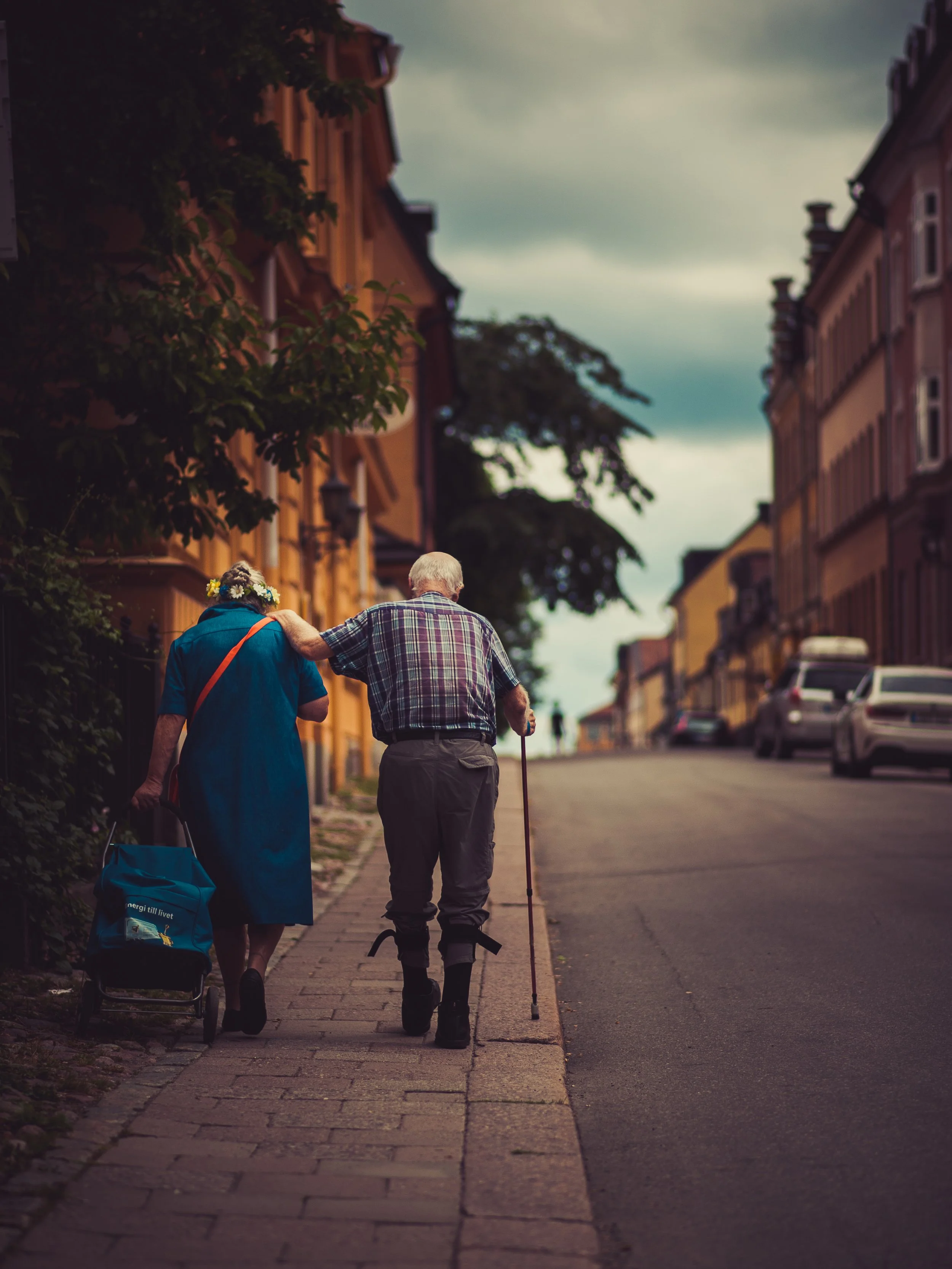 An elderly man with a cane walks with an elderly woman with a flower crown, helping her along a city sidewalk during late afternoon or early evening.