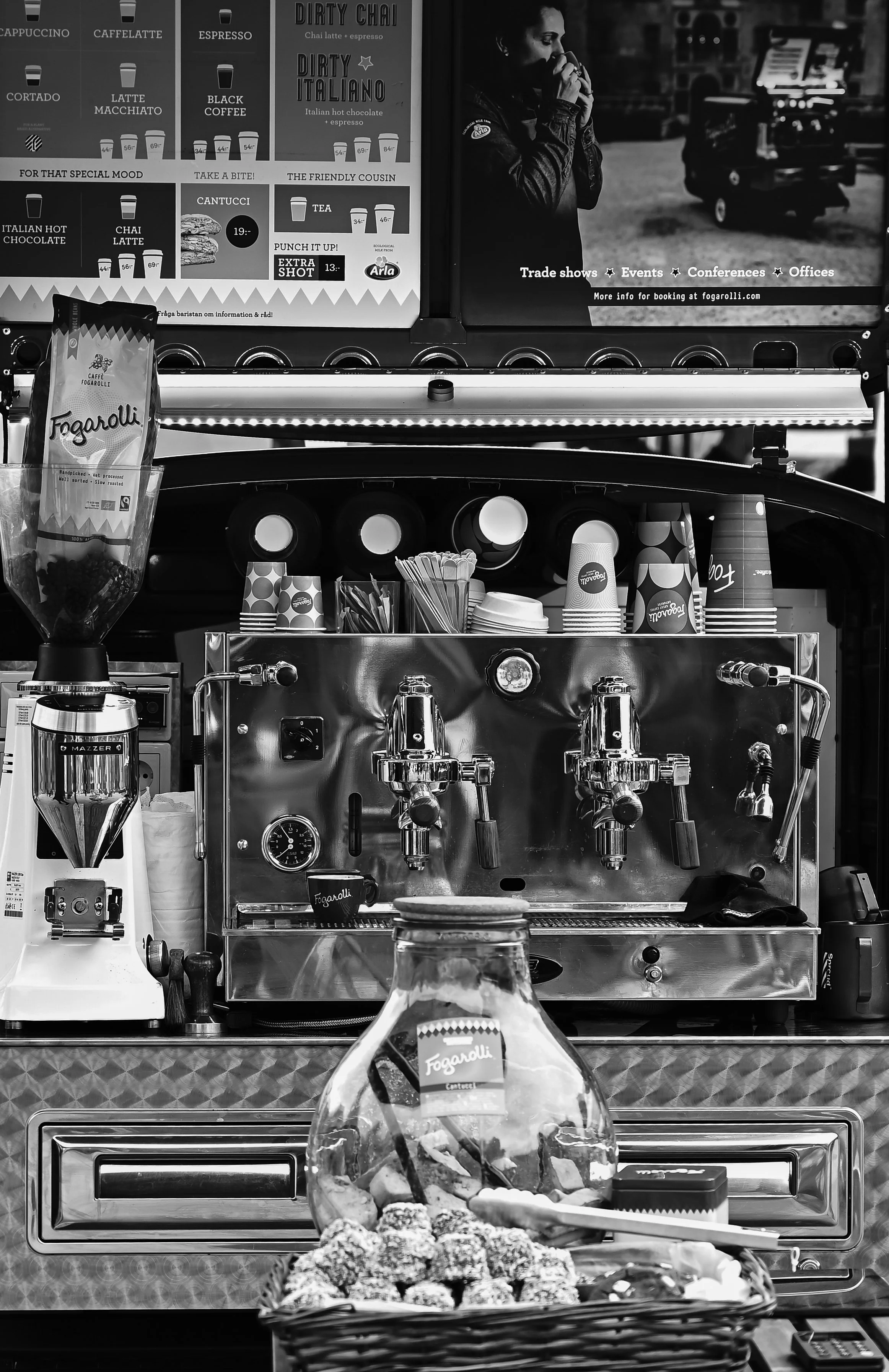A coffee stand with espresso machine, cups, stirrers, and a jar labeled 'Fogarolli' in front, with a menu above and a woman drinking coffee nearby.