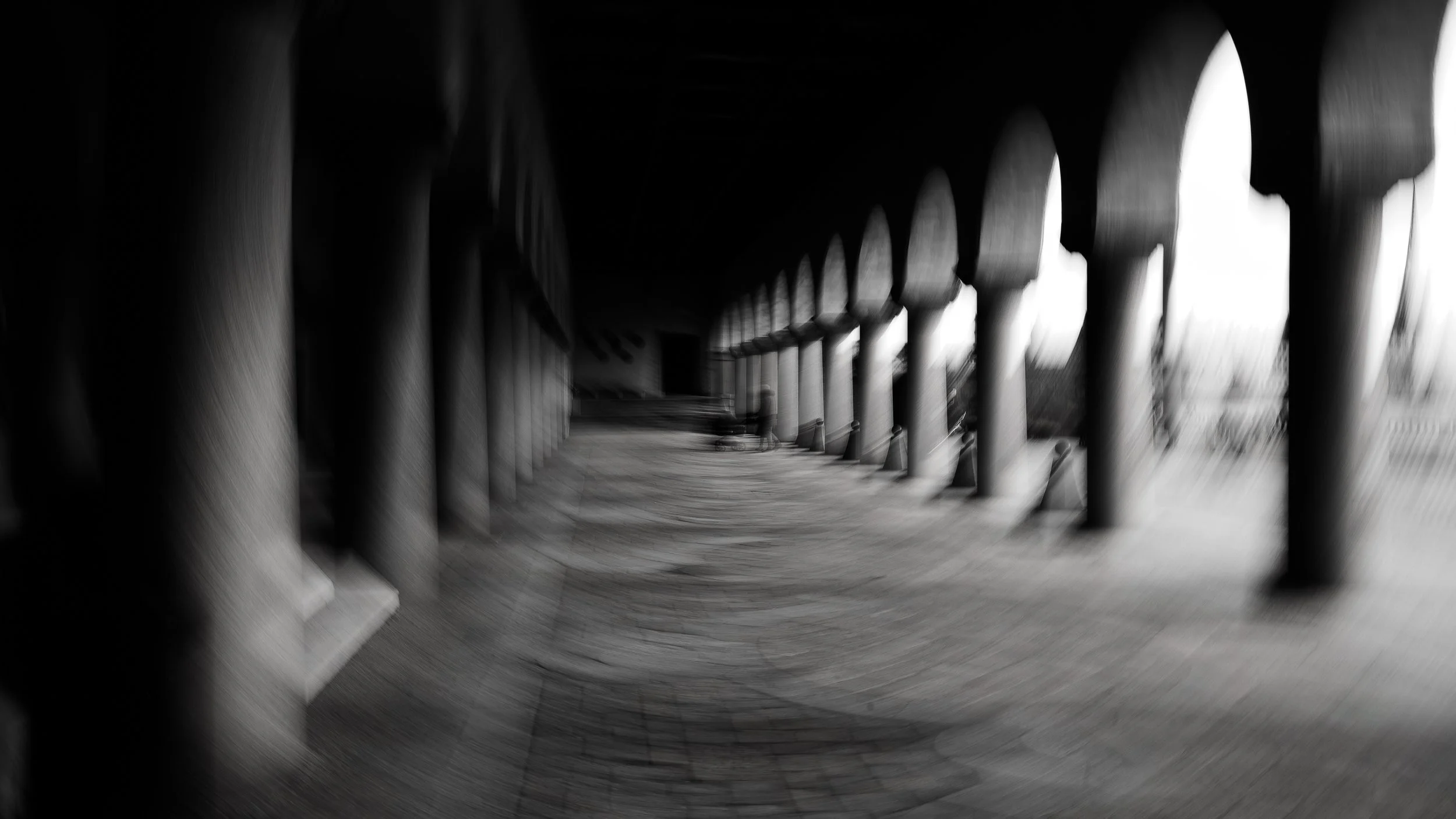 Black and white photograph of a covered outdoor walkway with arches and columns, with a curved pavement and a distant view of the outside.