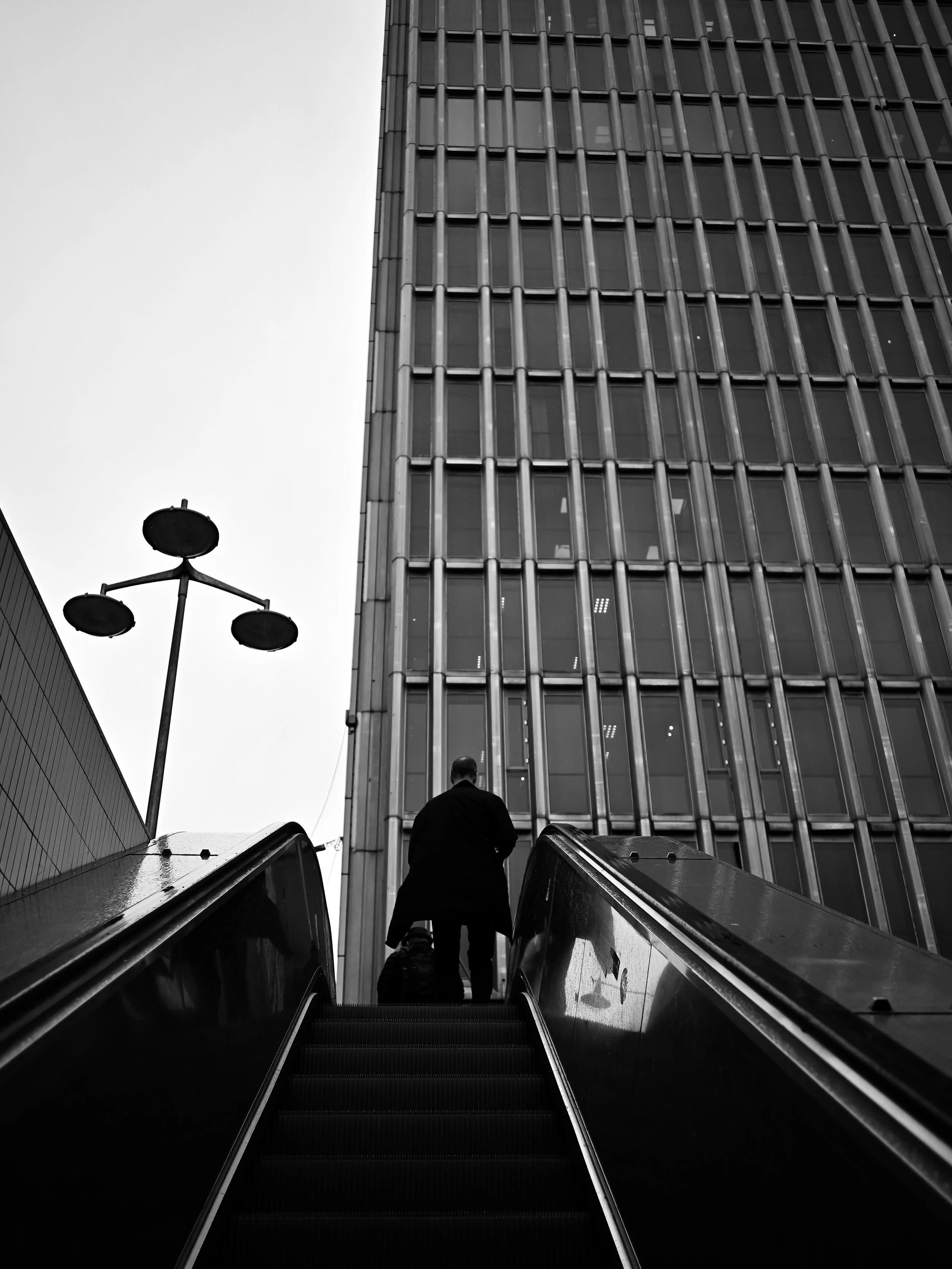 A person ascending an escalator towards a tall modern building with glass windows, with a streetlamp visible on the left, in a black and white photo.