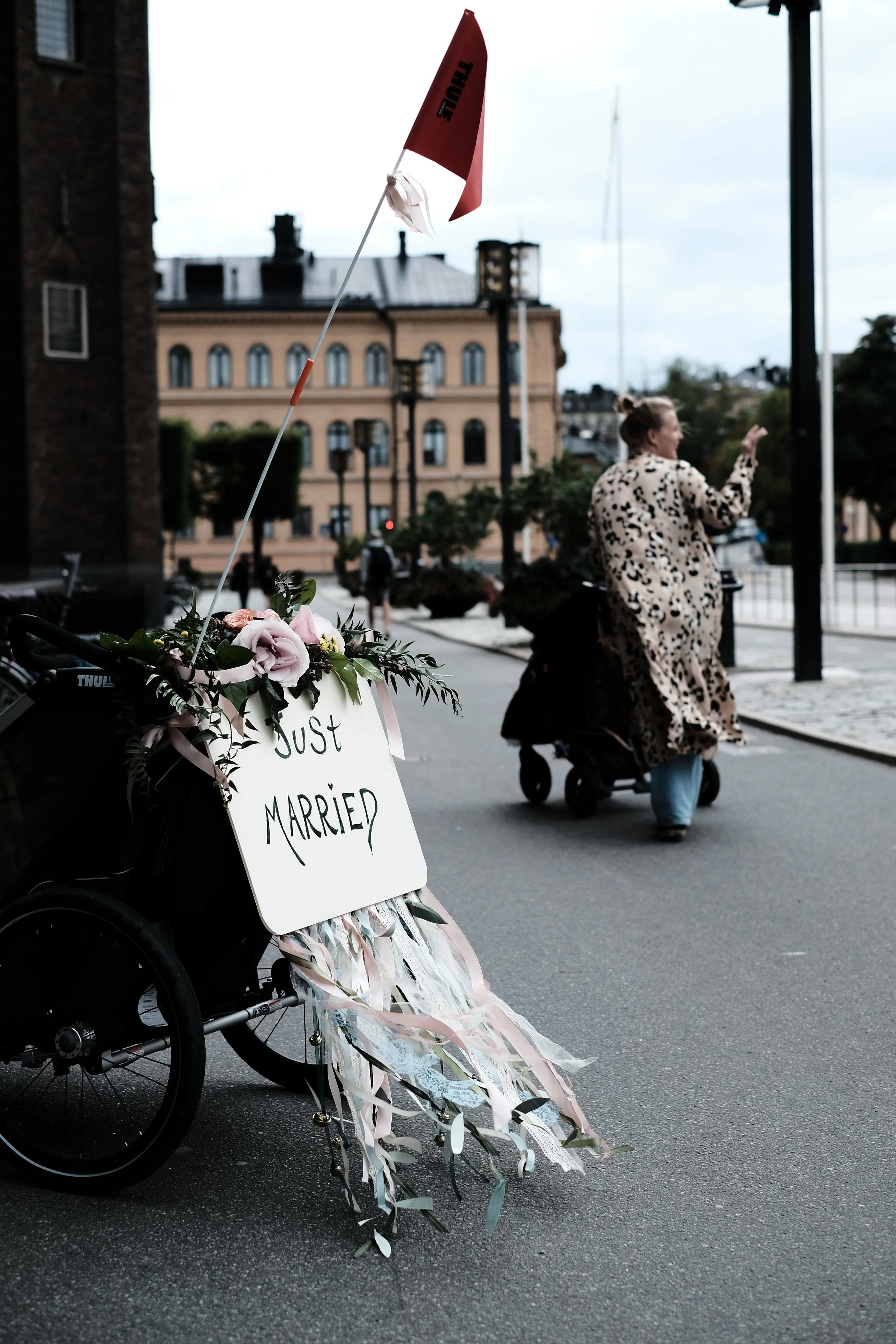 A wheelchair decorated with flowers and ribbons, with a sign that says 'Just Married,' parked on a city street during the day.