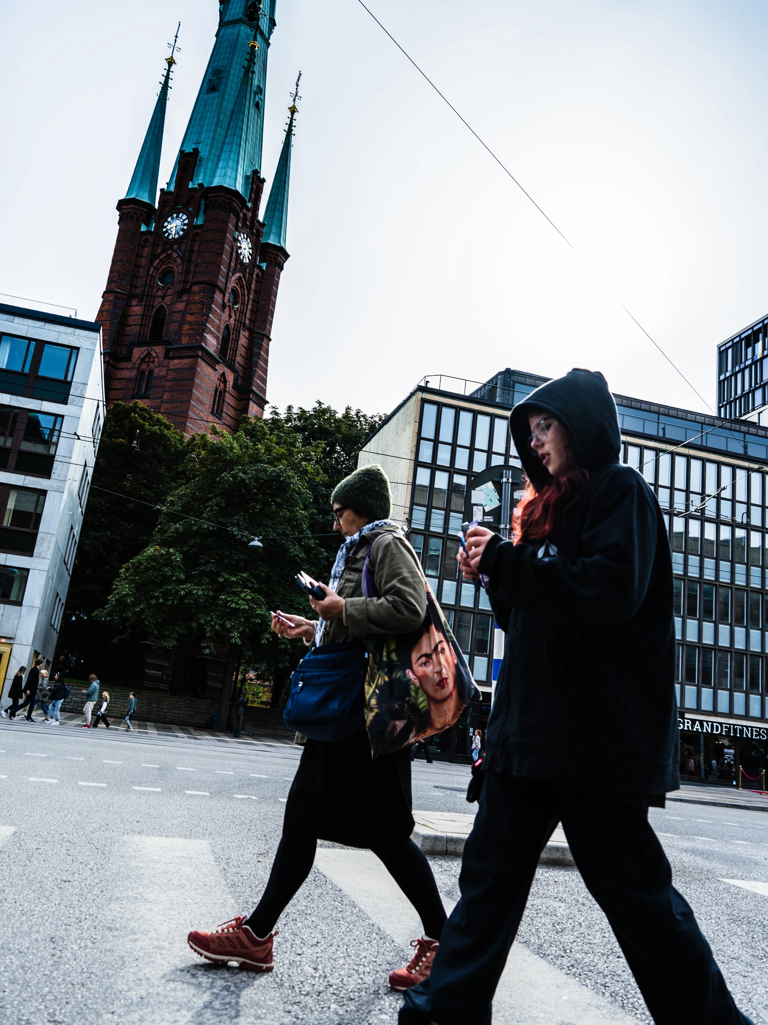 Two people walking on a city street, looking at their phones. One person is wearing a hooded jacket, glasses, and carries a tote bag with a woman's face printed on it. The other person is wearing a beanie, glasses, and a coat. In the background, ther