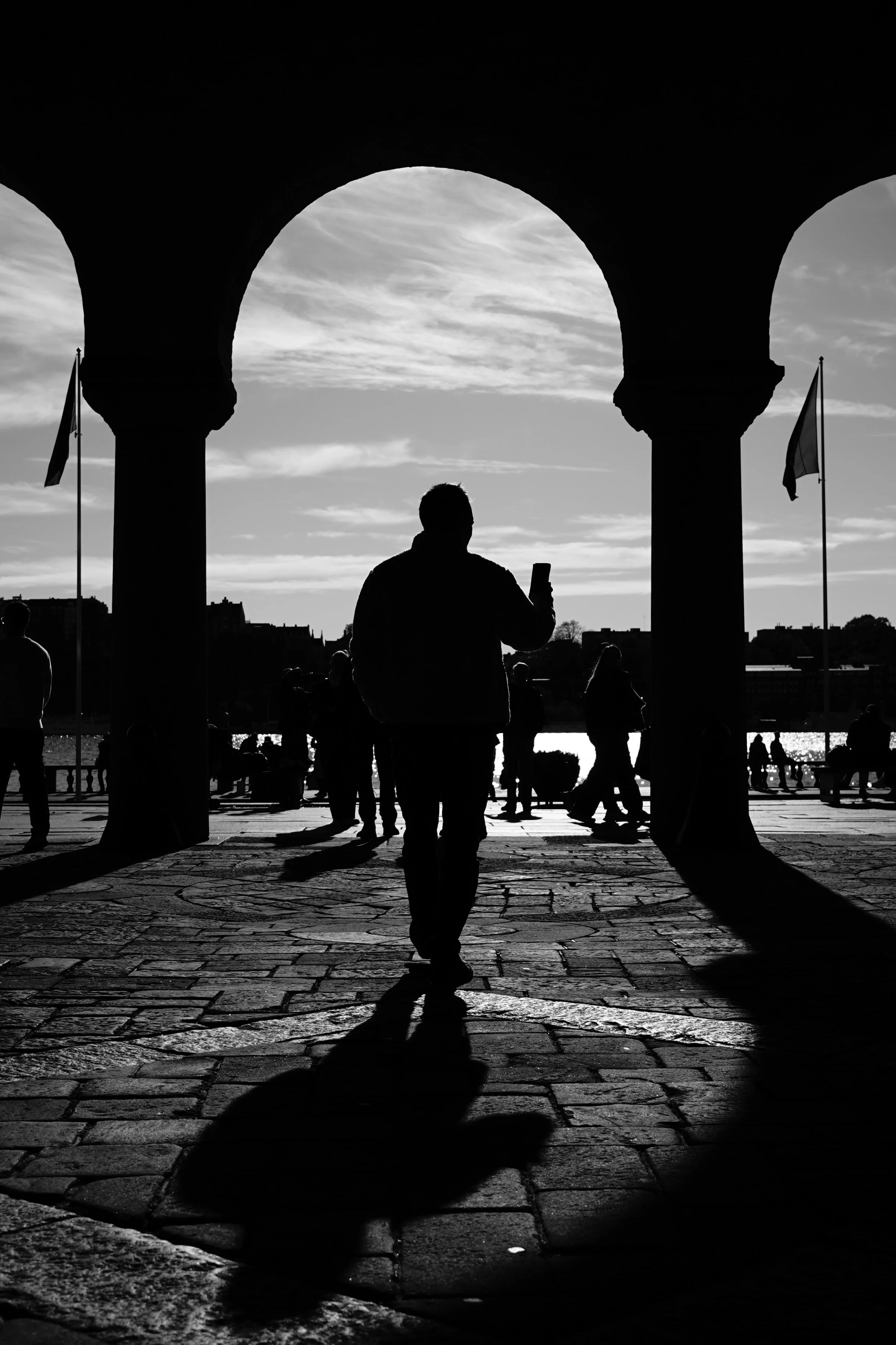 Silhouette of a man taking a photo on his phone under a large archway at sunset, with other people in the background near water and flags.