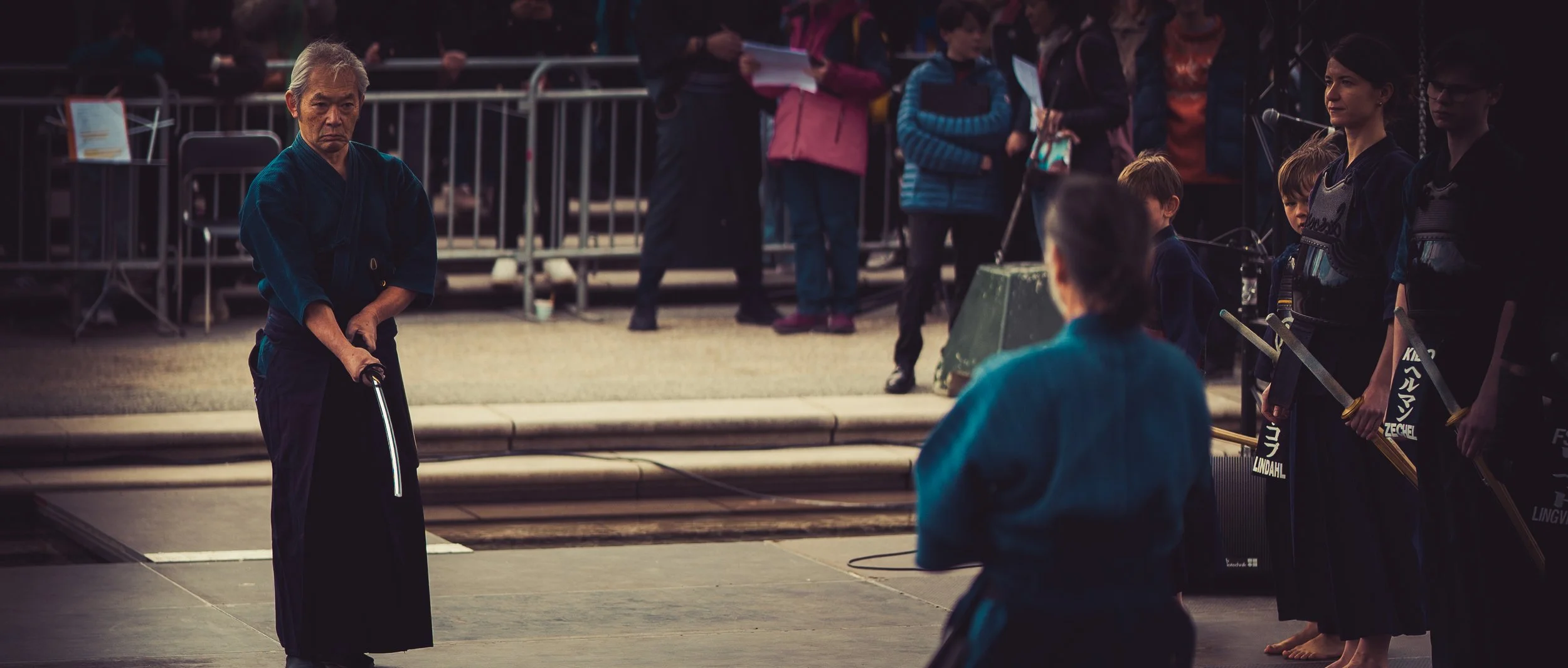A martial arts demonstration featuring an elderly man with gray hair holding a sword, surrounded by children and adults in martial arts uniforms and casual clothing watching outdoors, with metal barricades and onlookers in the background.