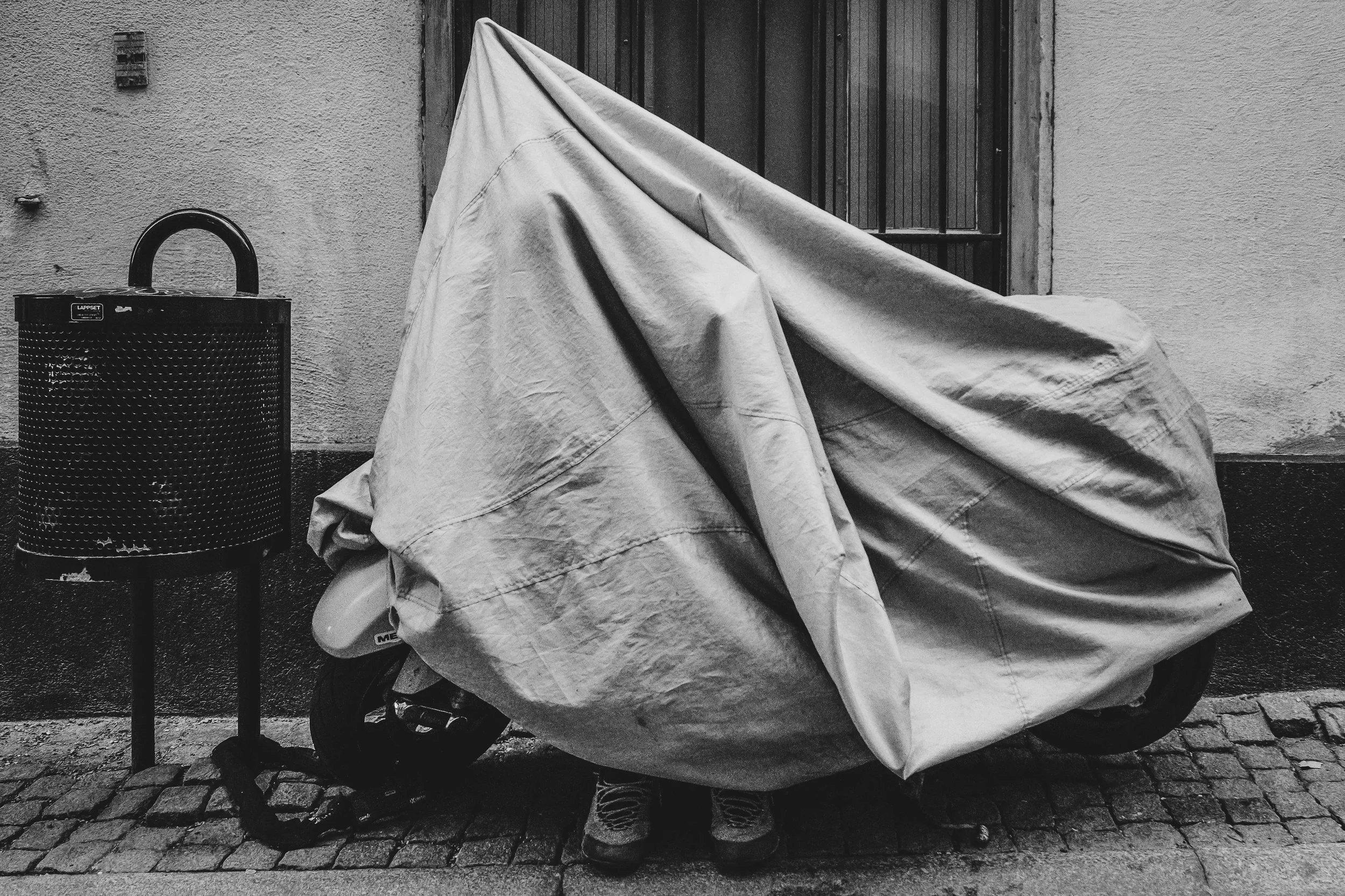 A person with shoes visible beneath a large, draped cover leaning against a wall on a cobblestone sidewalk, next to a trash can and a window with bars.