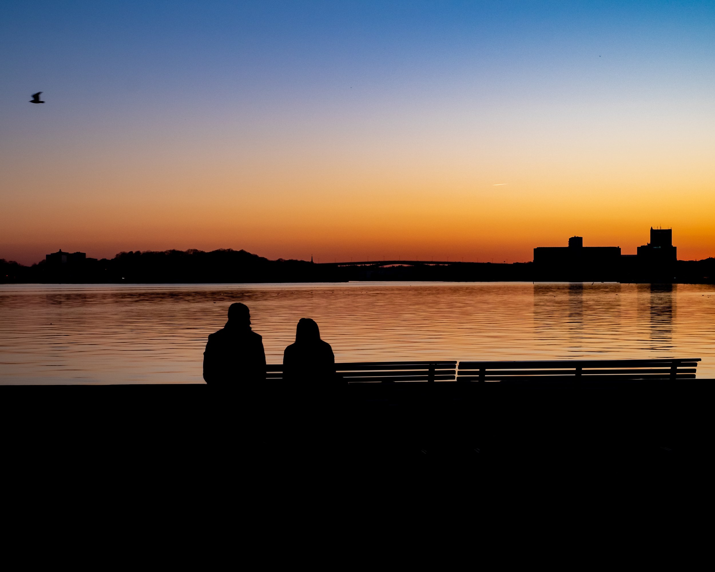 Silhouettes of two people sitting on a bench near a body of water during sunset, with a colorful sky and city skyline in the background.