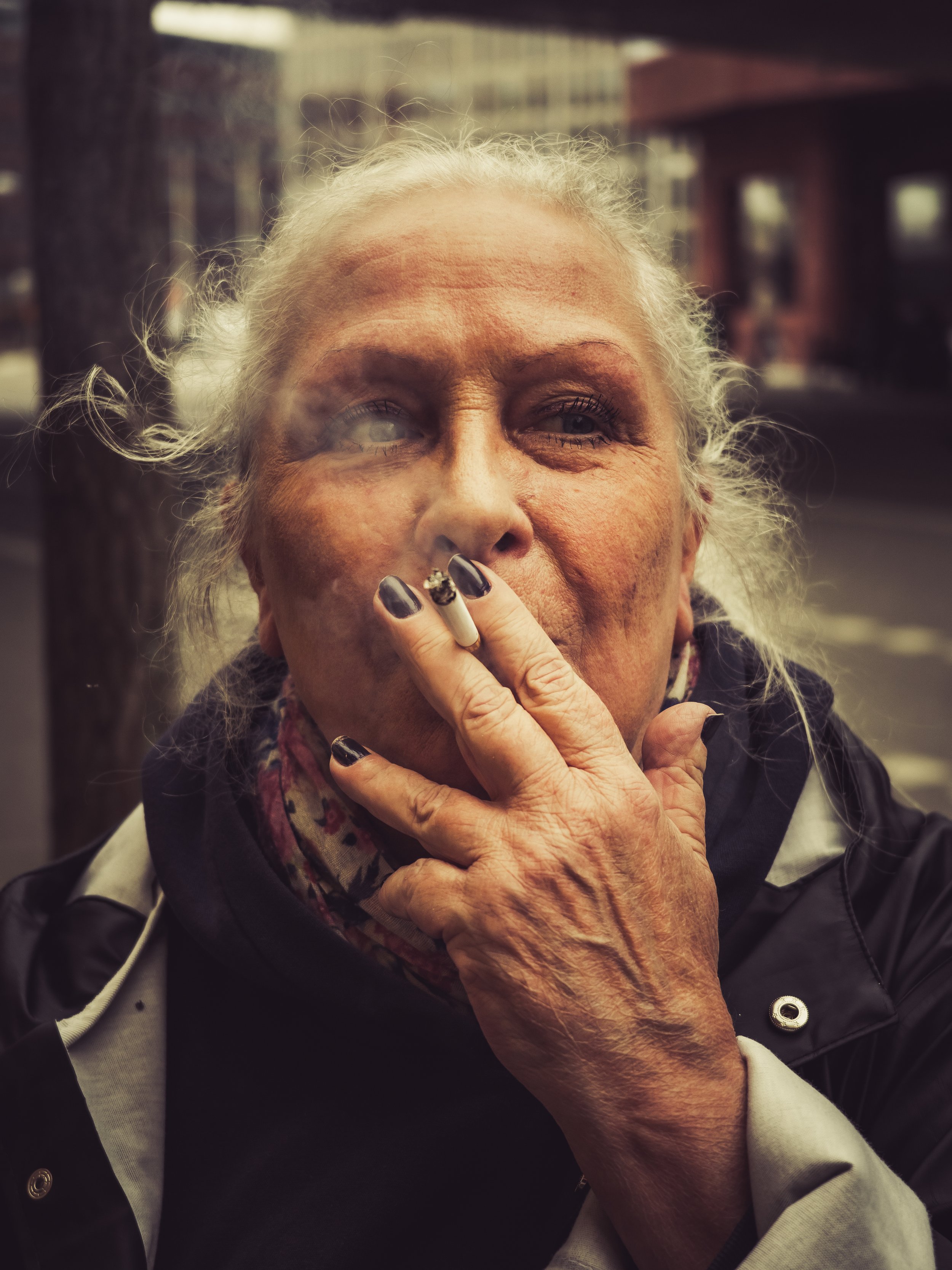 Older woman with gray hair smoking a cigarette outside on a city street.