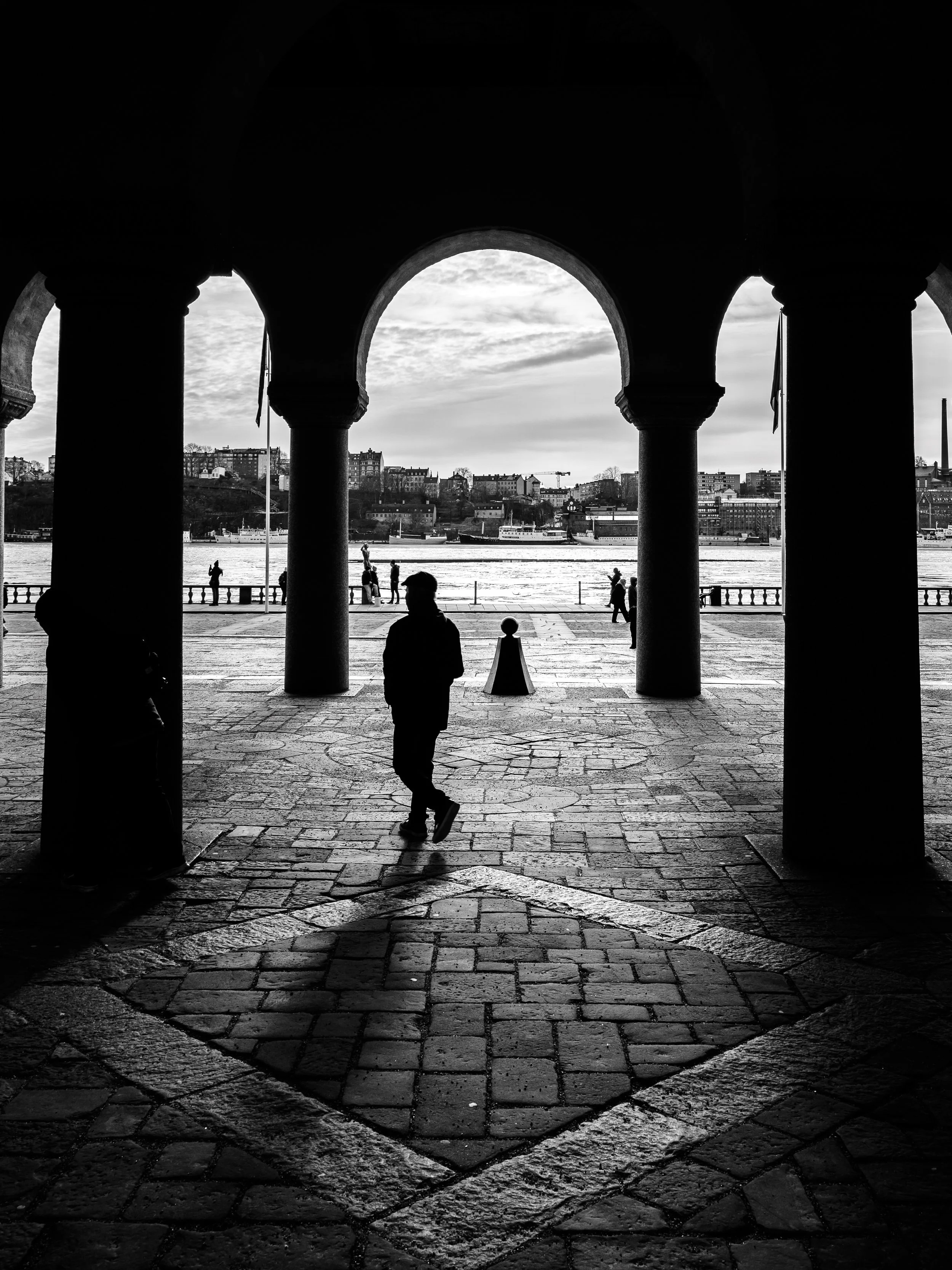 Black-and-white photo of a person walking under arches near a body of water with buildings in the background.