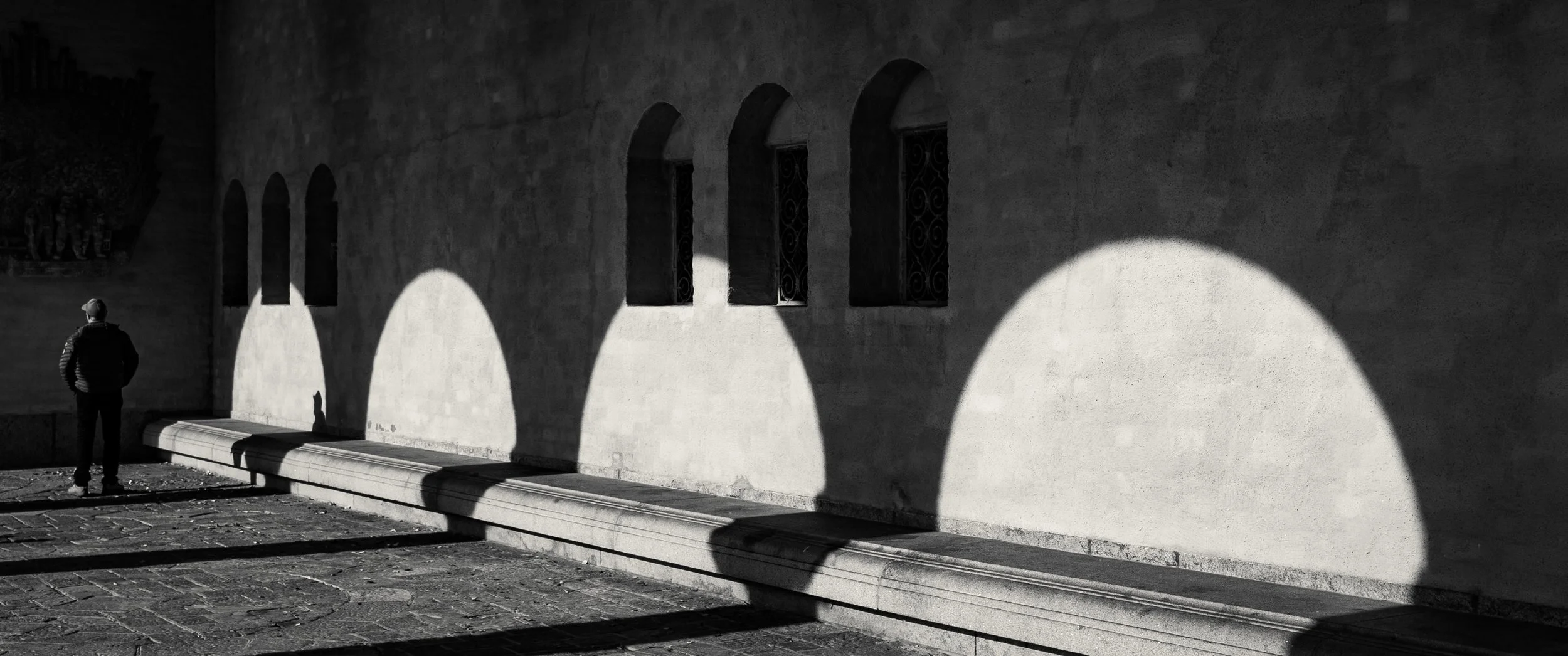 A black and white photo of a man standing on a cobblestone street near a stone wall with three arched windows. Shadows cast on the wall create large circular shapes.