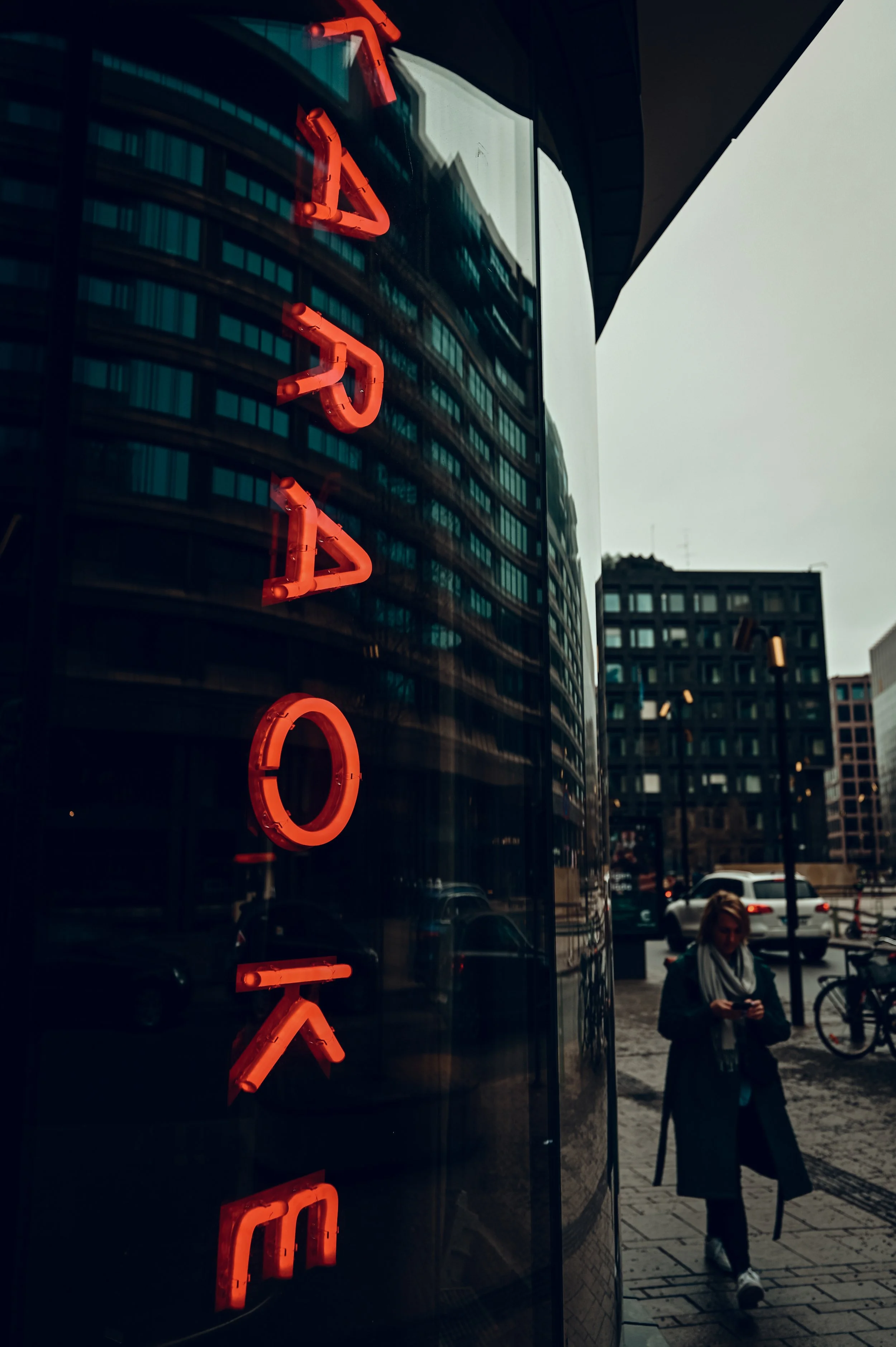 Reflection of city buildings on a glass window with a red neon sign that says 'LOVE', and a woman walking on the sidewalk using her phone.