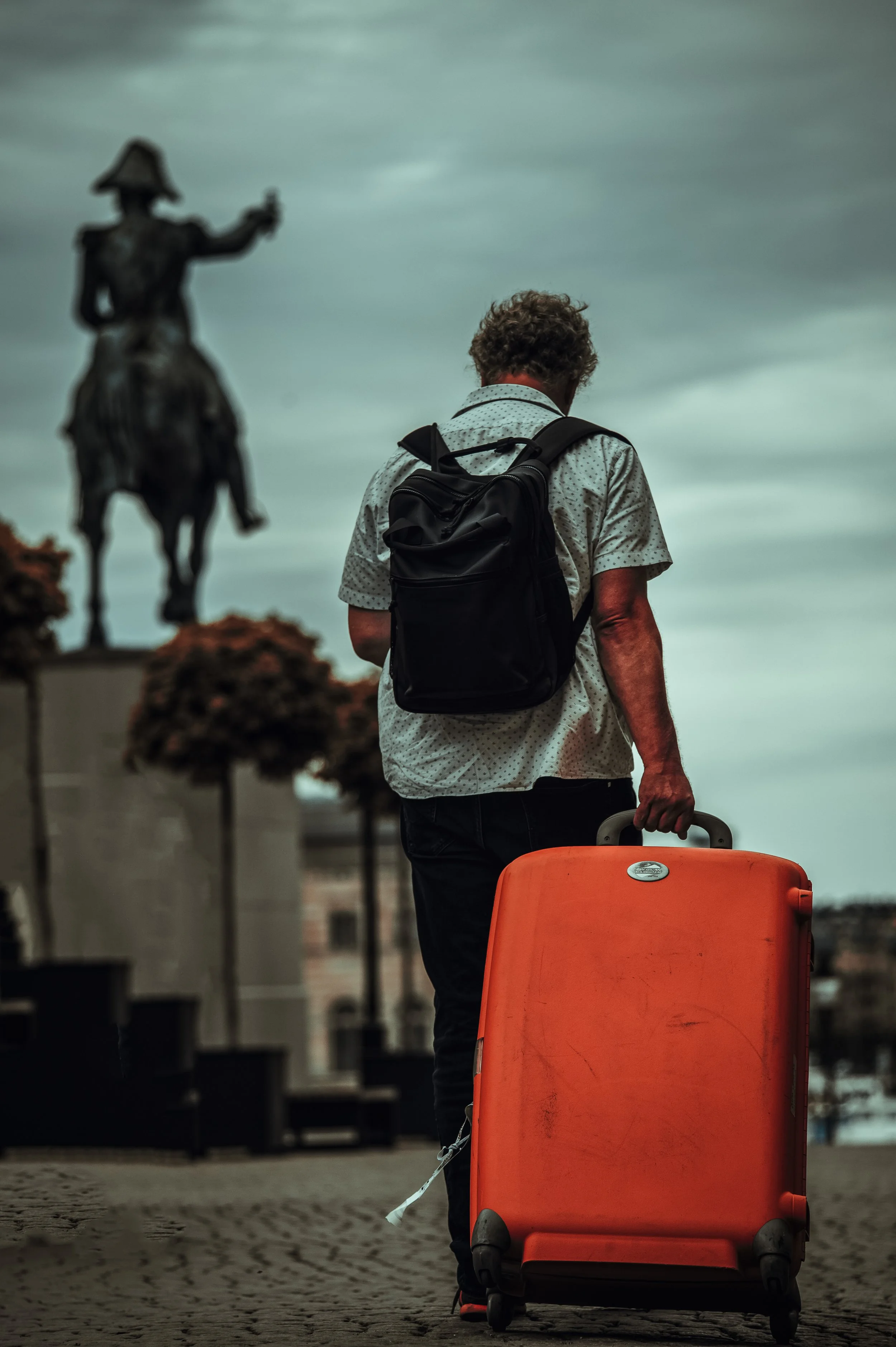 A man with gray curly hair, wearing a white shirt with small black dots and a black backpack, pulls an orange suitcase while walking in an outdoor area with a statue on horseback and trees in the background.