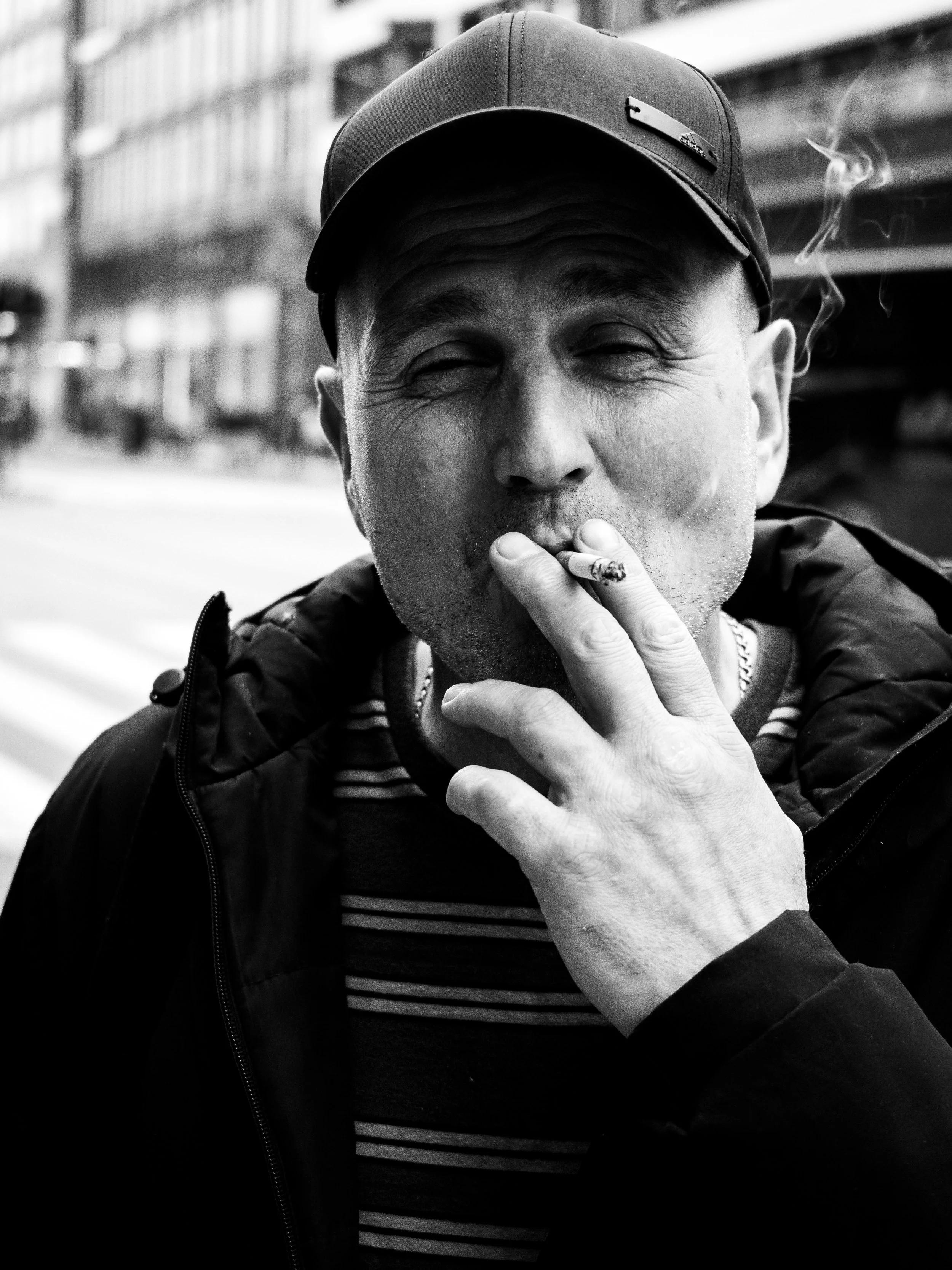 Black and white photo of a man smoking a cigarette outdoors, wearing a cap and a jacket, with buildings in the background.
