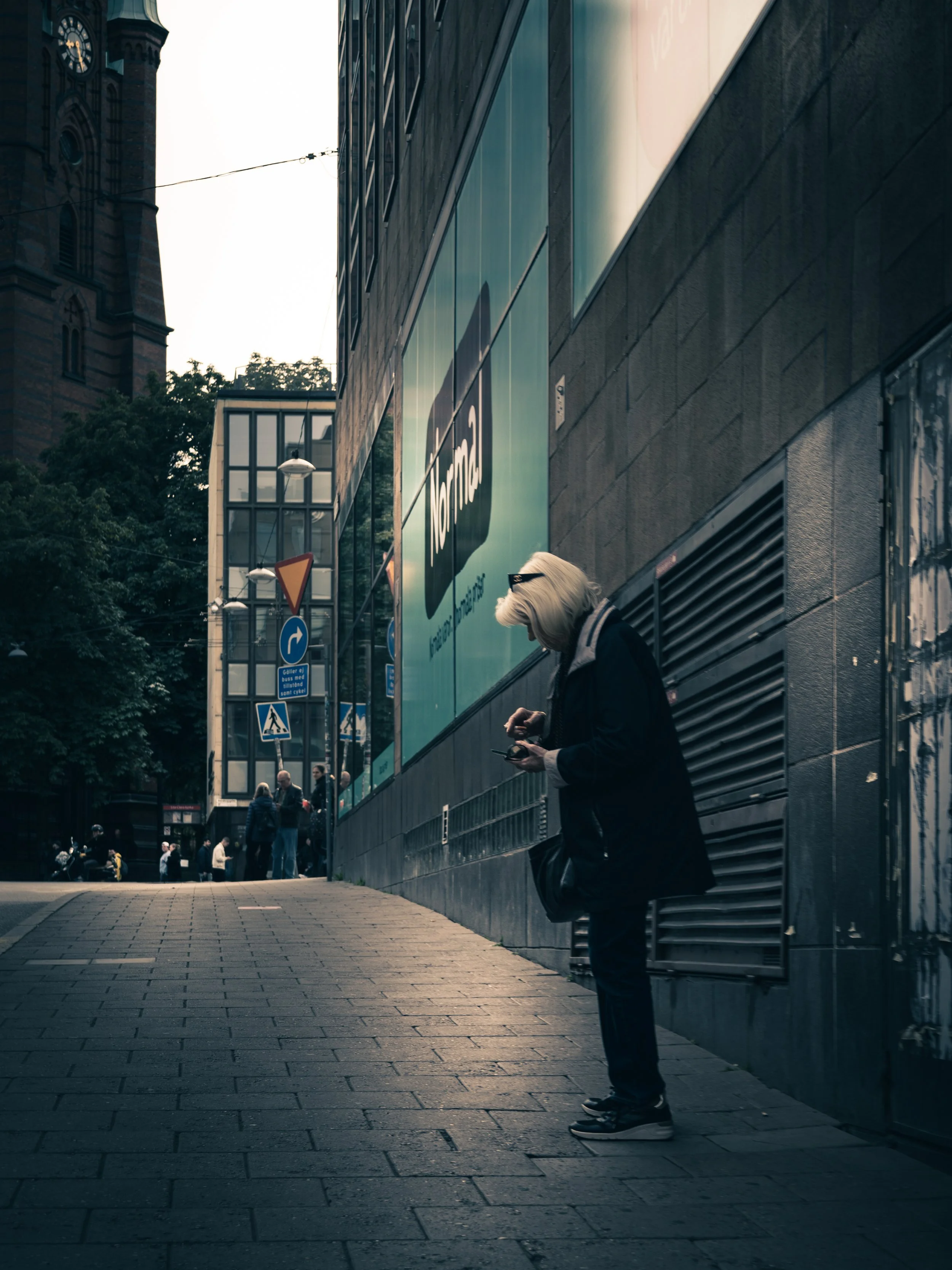 A person with blonde hair, wearing dark clothing and glasses, standing on a city sidewalk, looking at their phone, with a large building and traffic signs in the background.