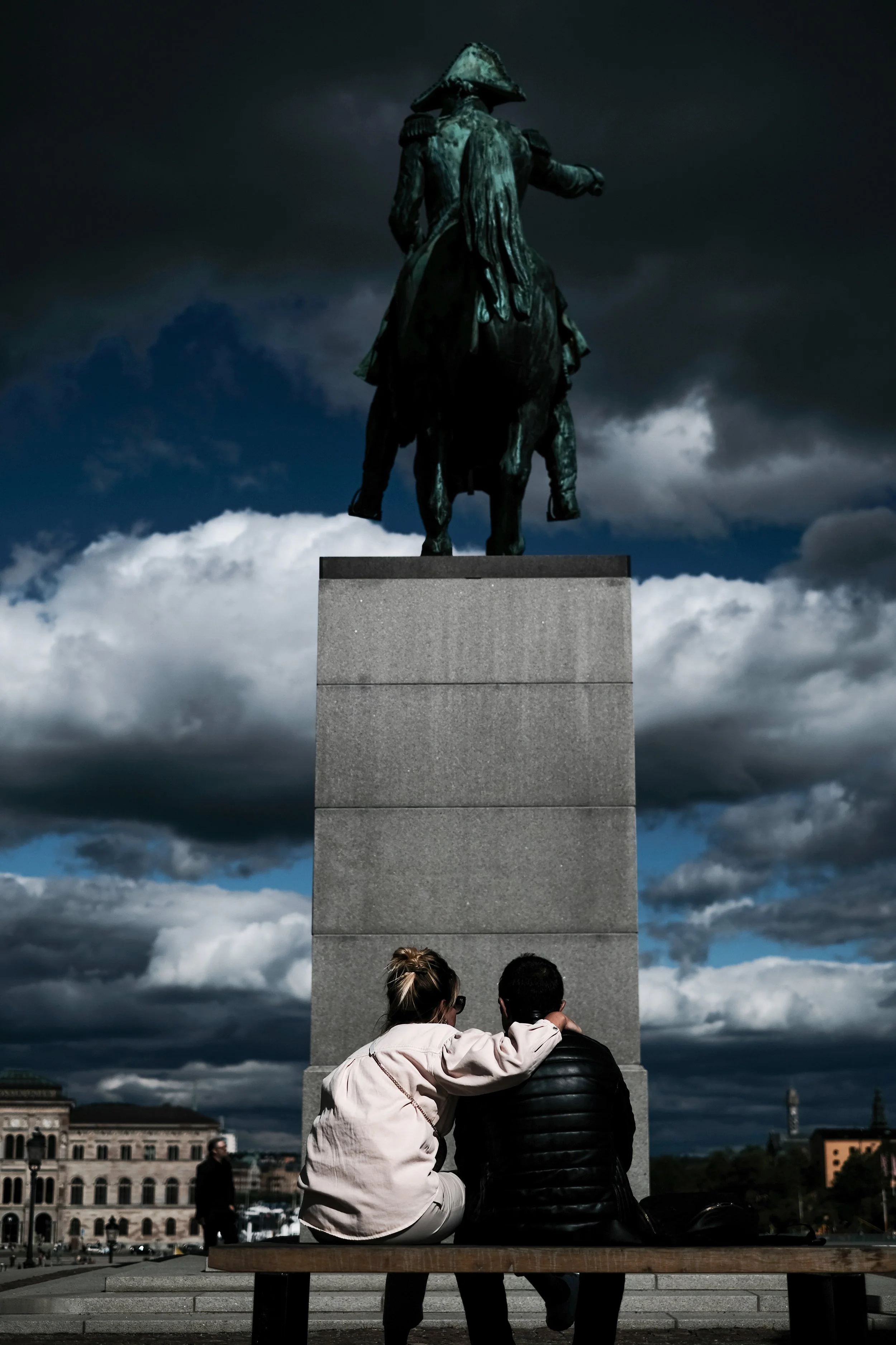 A man and woman sitting on a bench, with the woman leaning on the man's shoulder, looking at a large equestrian statue of a person in historical clothing and a hat, under a cloudy sky.