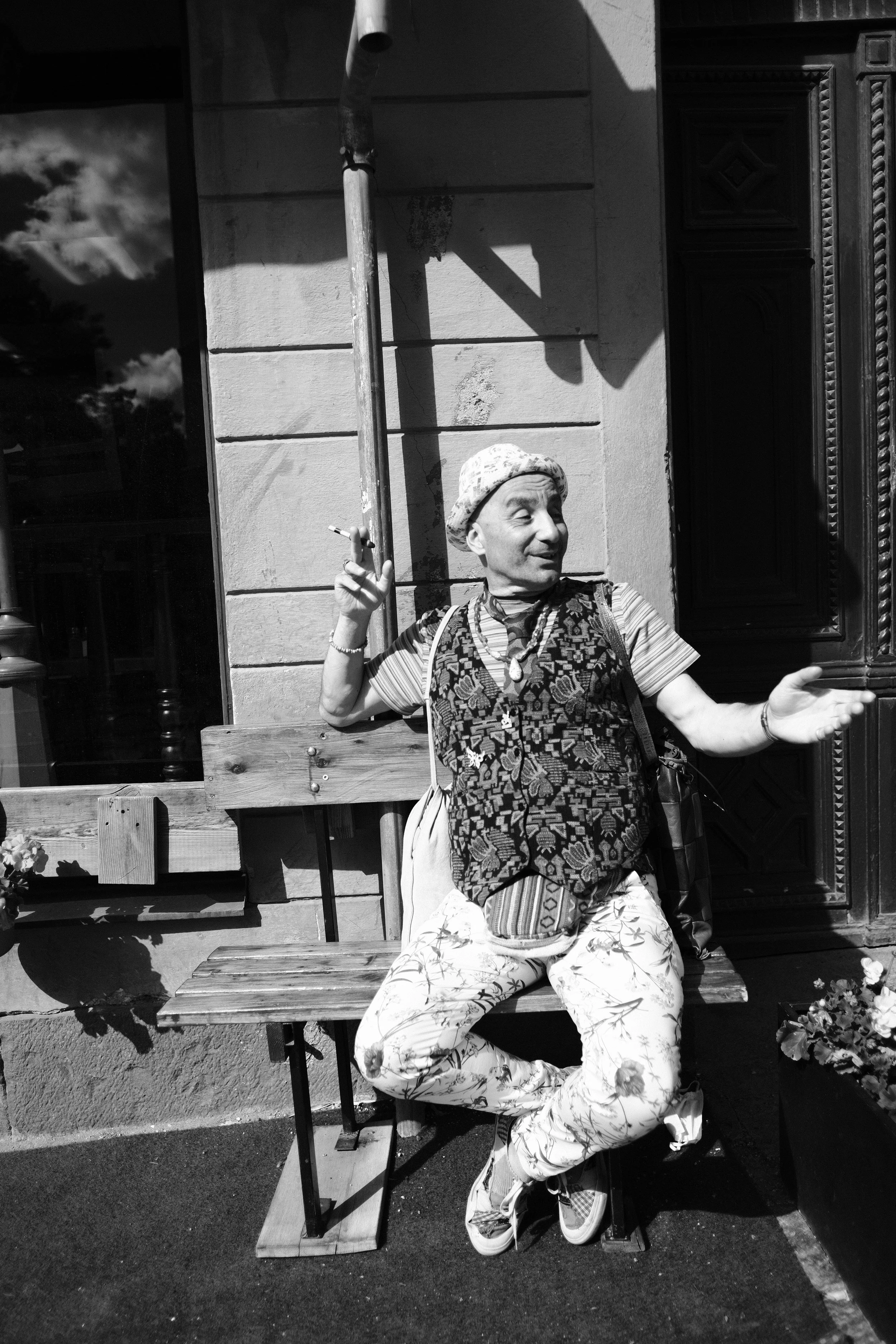 A black and white photo of a person sitting on a wooden bench outside a building, wearing a patterned vest, floral pants, a hat, and holding a cigarette. The person is gesturing with one hand, with a playful expression.