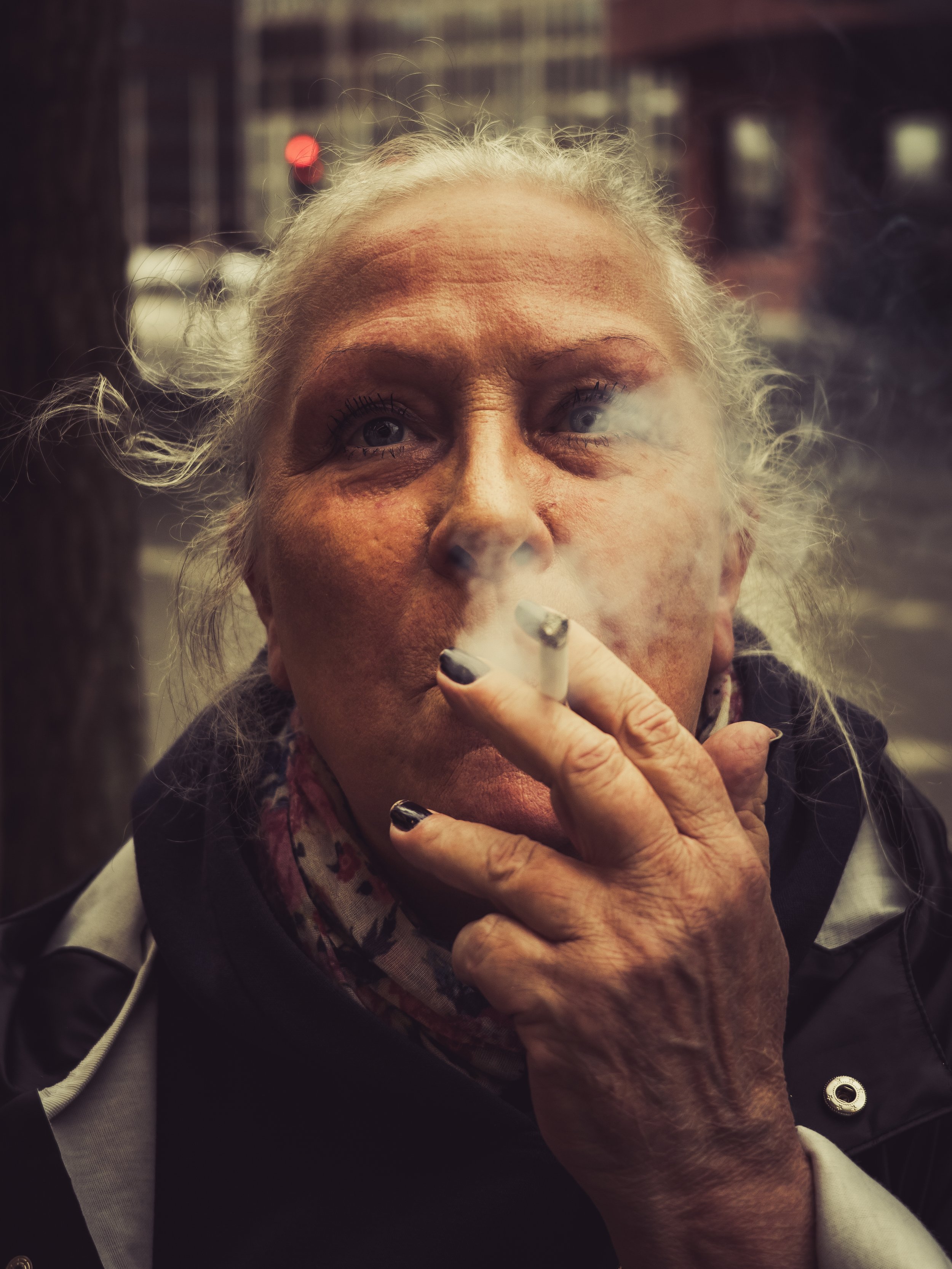 An elderly woman with gray hair and blue eyes smoking a cigarette outdoors, with smoke visible and city buildings in the background.