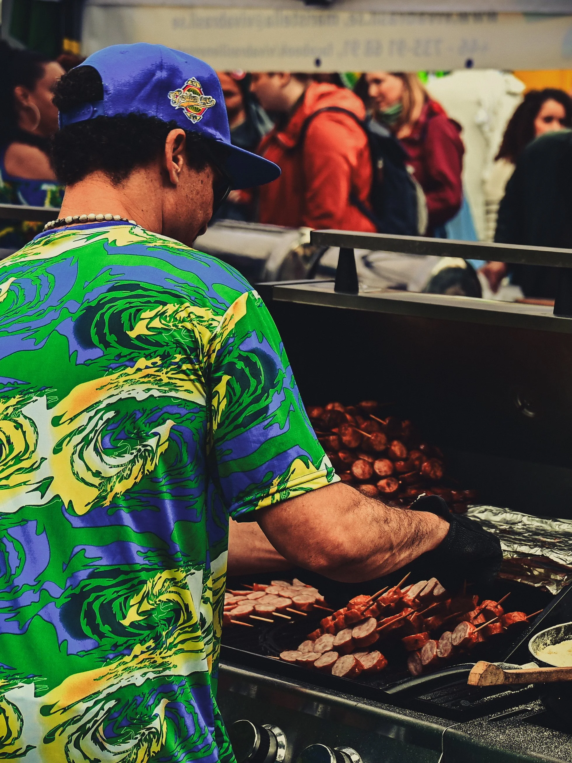 A man grilling skewered sausages at a busy outdoor food stall on a sunny day, with a crowd of people in the background.