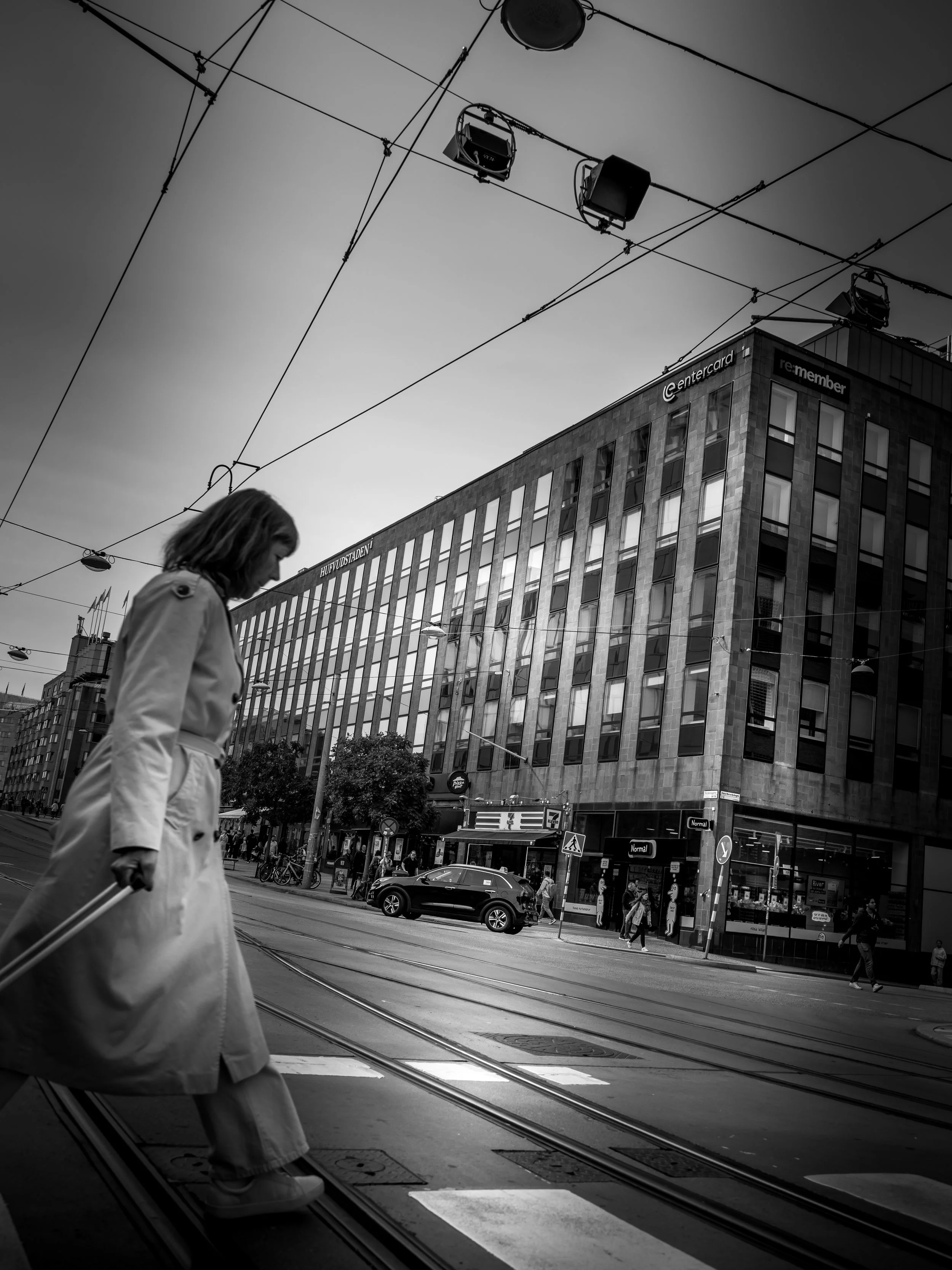 A woman crossing the street on a pedestrian crosswalk in an urban city with tram tracks, modern multi-story buildings, and overhead tram wires in black and white.