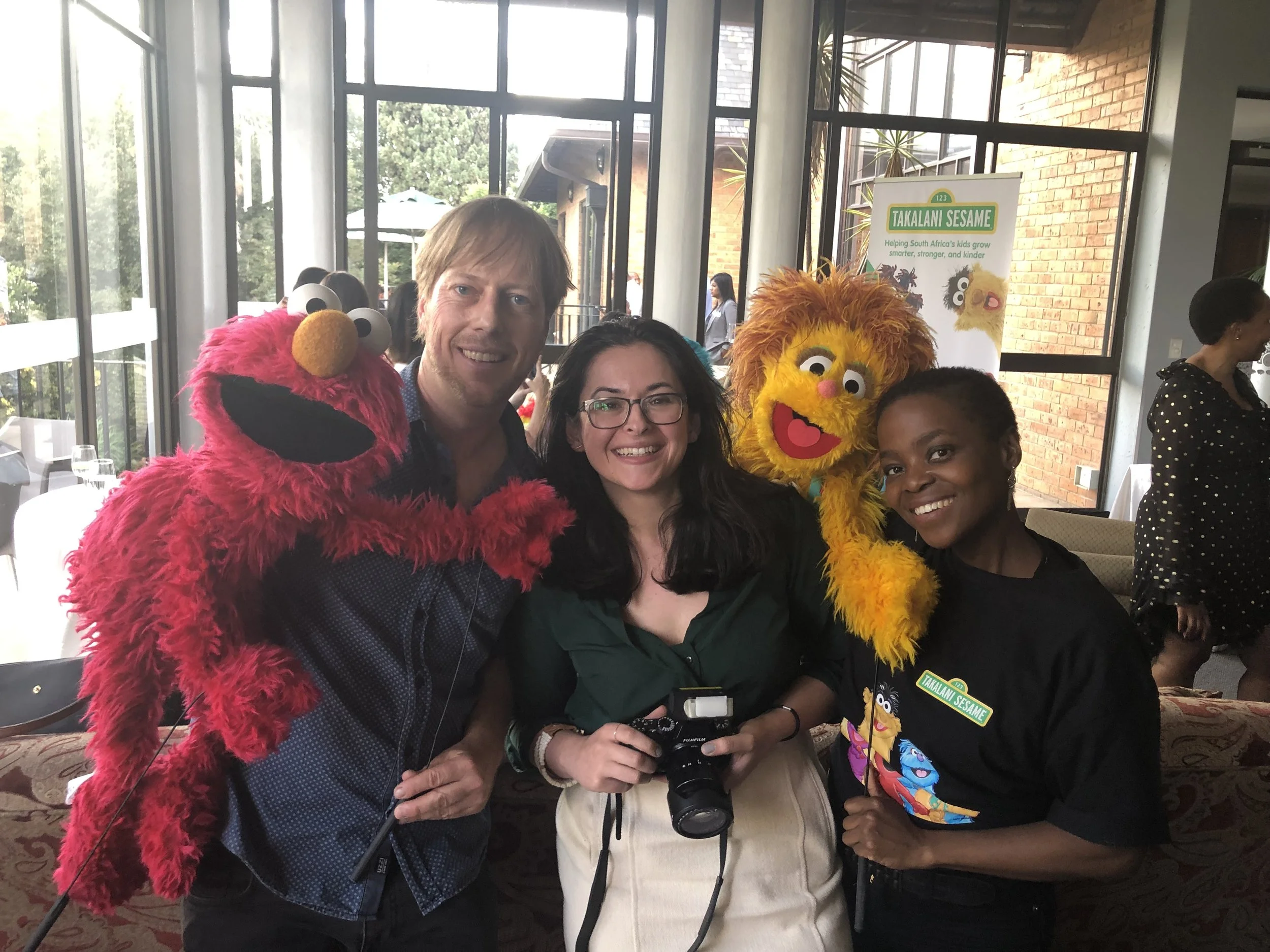 Bettina (in the middle with a camera) poses with two puppateers and their puppets from the Takalani Sesame production. The two puppets are Neno (Elmo for US audiences) and Kami (South Africa’s HIV-positive muppet).