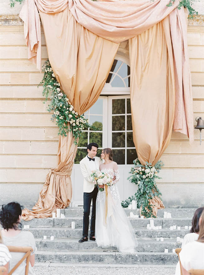 A bride and groom standing on stairs with flowers, candles and canopy at the front of a French chateau wedding venue.