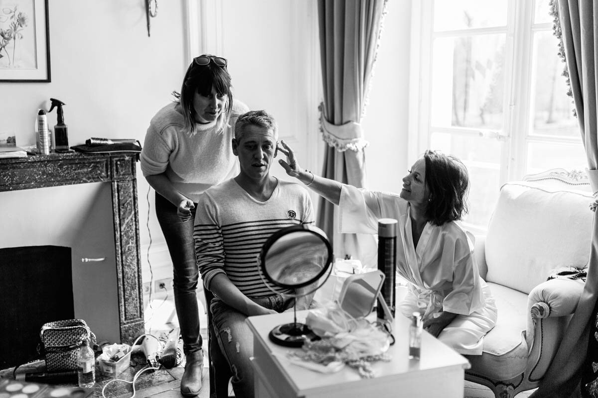 A black and white photo of a bride getting ready with a makeup artist and a man, at a French destination wedding in a chateau.
