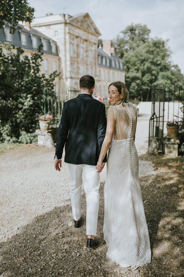 A bride with her head turned to the camera smiling and groom walking down a path in front of Chateau de Courtomer at an intimate wedding.