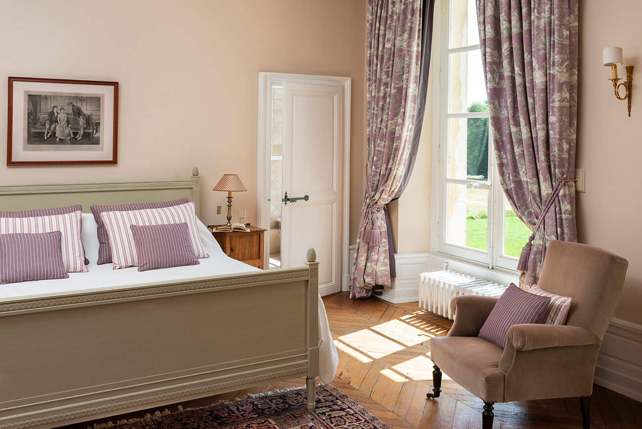 A light and airey bedroom with a double bed and a lounge chair with great views available to rent in a French chateau.