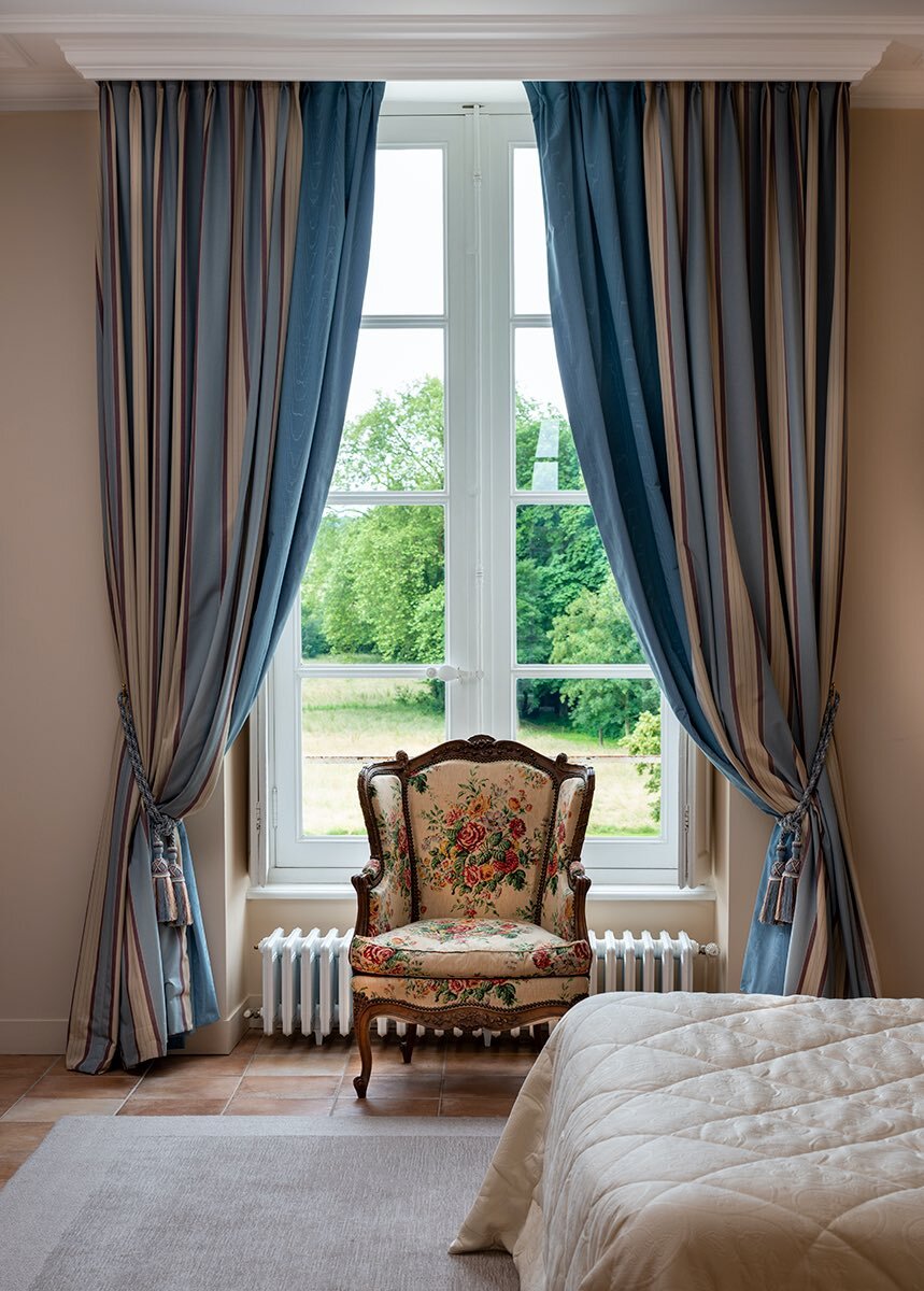 A bedroom with blue curtains and antique chair in front of a window, perfect for those looking to rent a chateau in France.