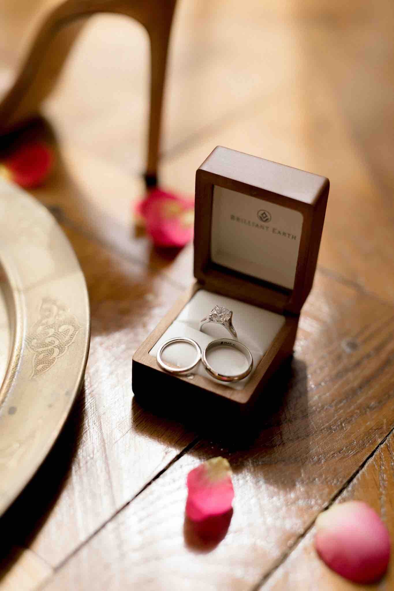 Two wedding rings side-by-side in a box on a wooden table with rose petals and a wedding shoe.