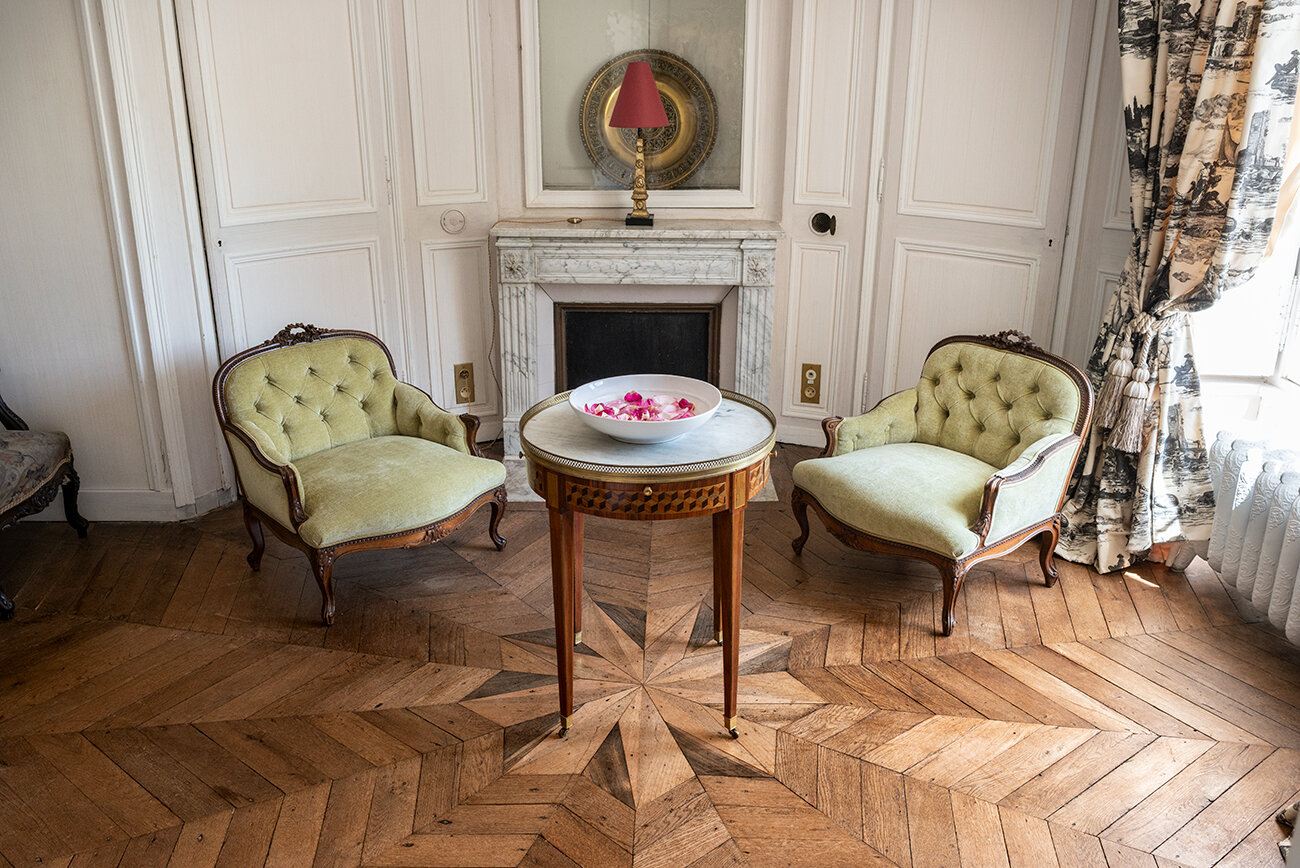 A room with two antique chairs and a cozy fireplace in a chateau near Paris available for rent.