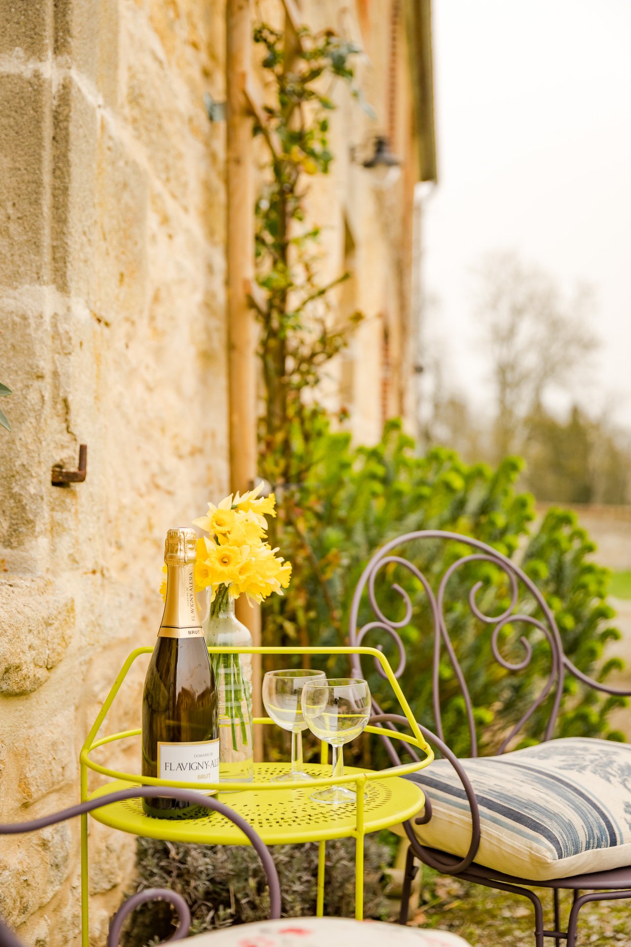 A bright yellow garden table with wine glasses and a bottle of wine on it, on the terrace of a  holiday cottage in Normandy.