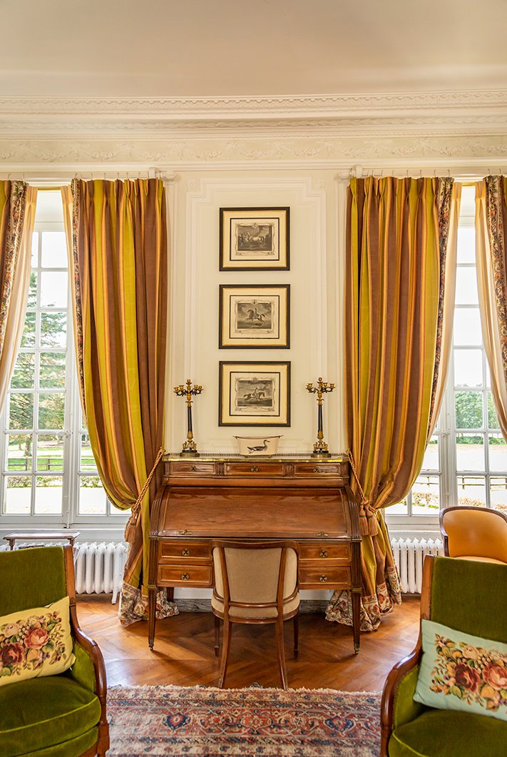 A living room with an antique bureau piano and occasional chairs, with grand windows and drapes in a wedding chateau near Paris.